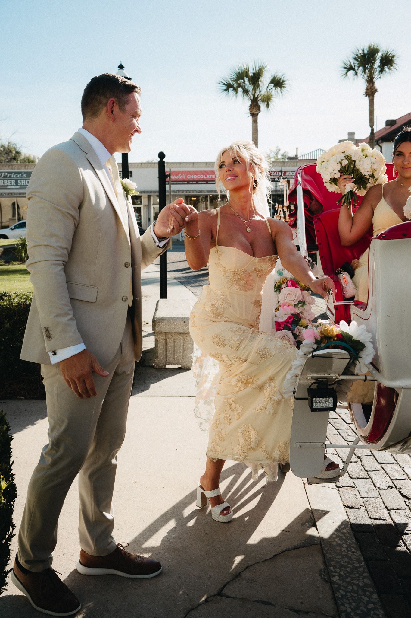 Bride in ivory tea-length dress holds bouquet while groom in tan suit holds her hand outside on sunny wedding day.