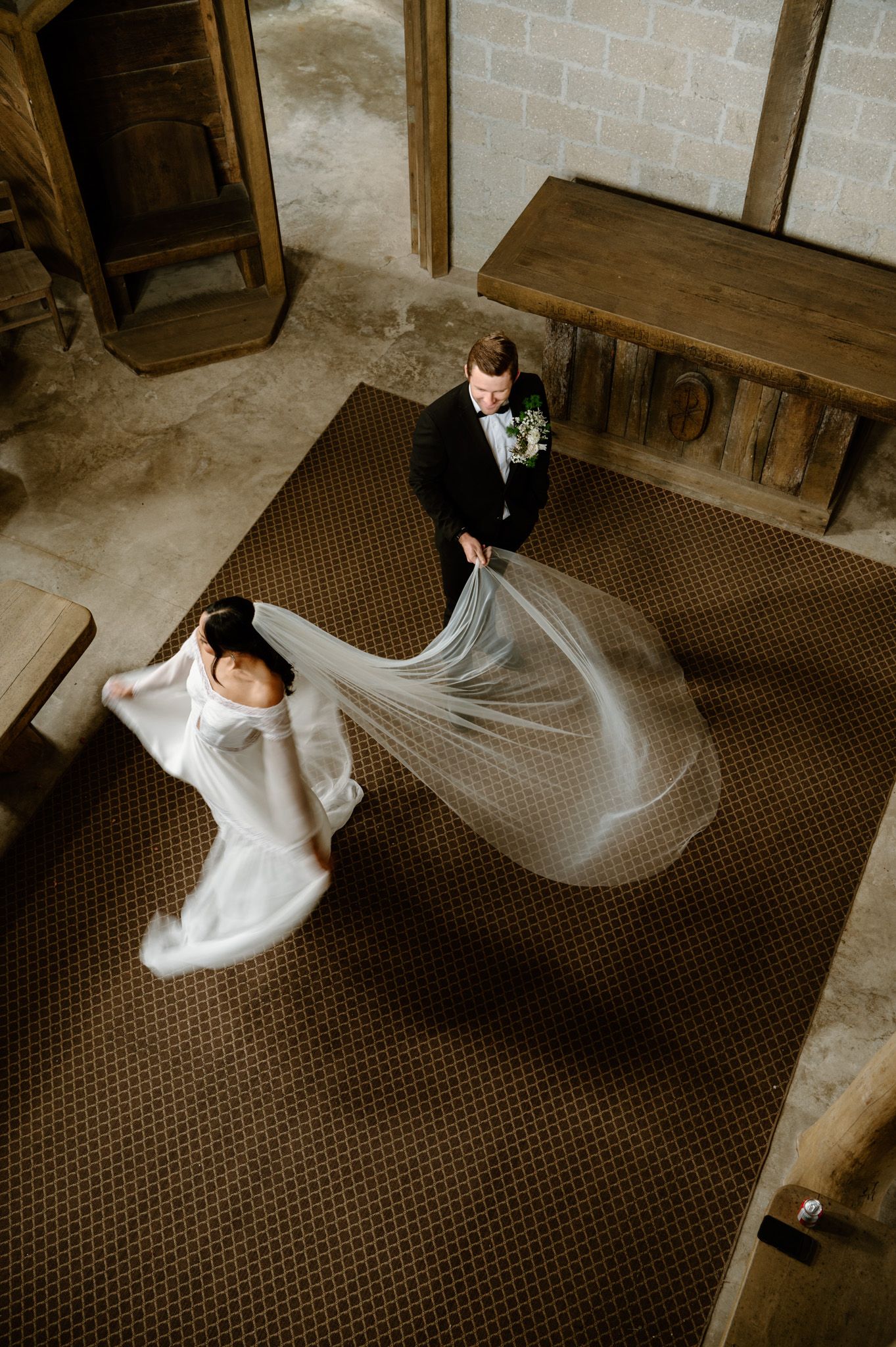 Aerial view of groom holding bride's flowing cathedral veil as they walk across a patterned floor inside a chapel.