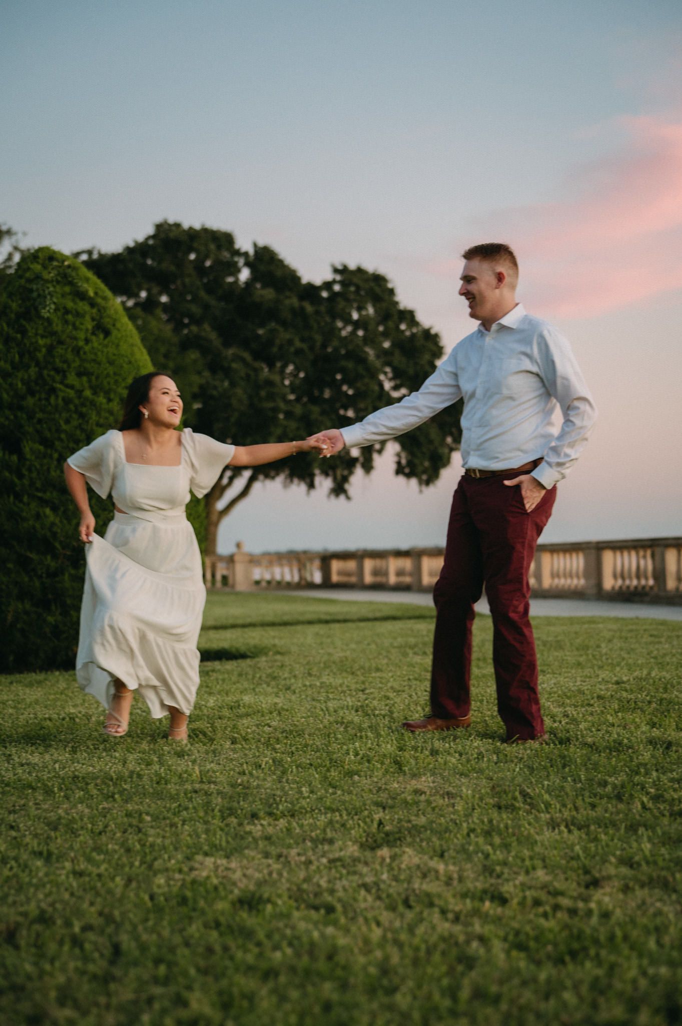 Joyful couple holding hands dancing on green lawn at sunset during engagement photo session.