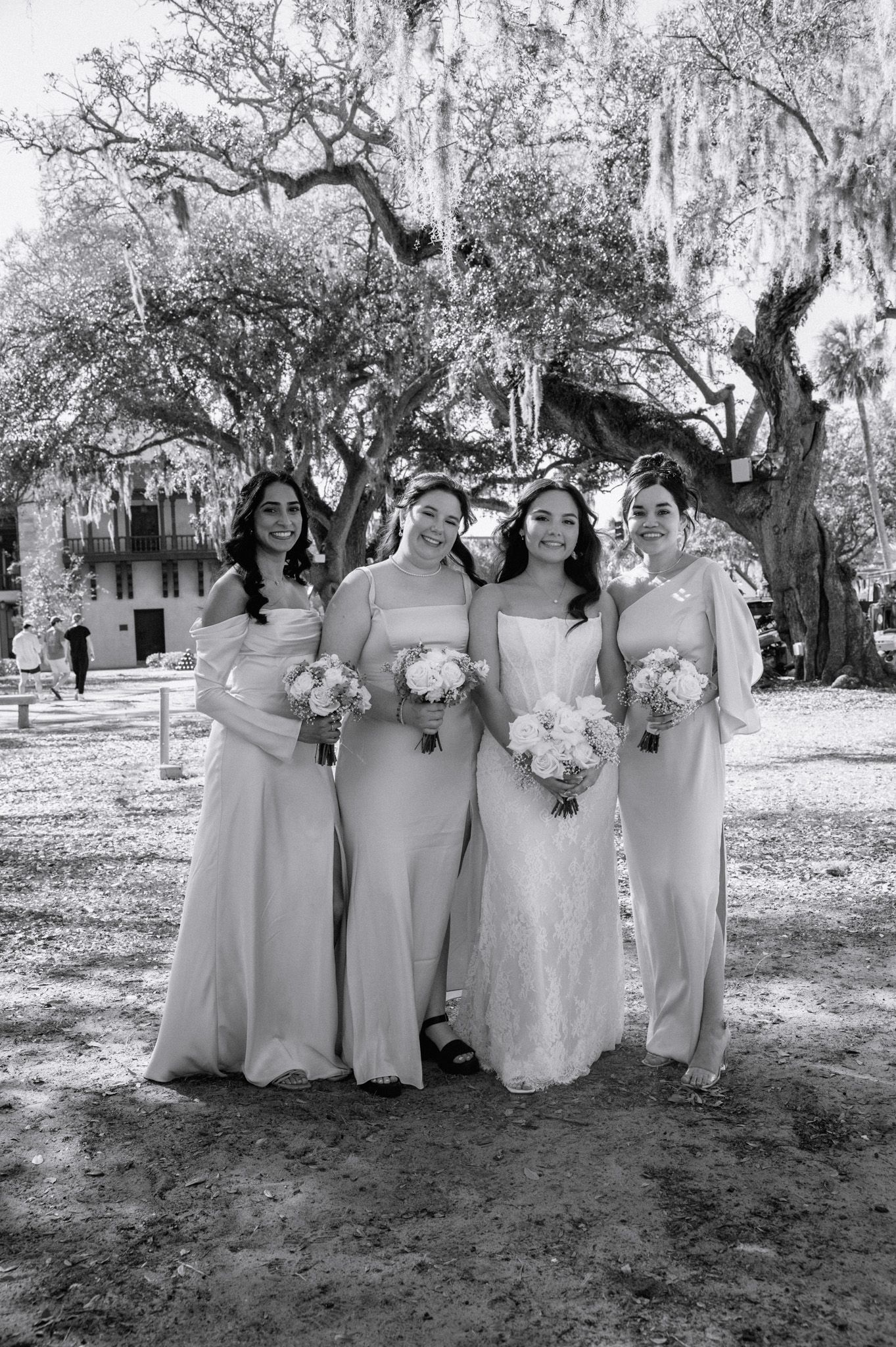 Black and white photo of a bride in lace gown with three bridesmaids in elegant dresses holding bouquets under a large tree.