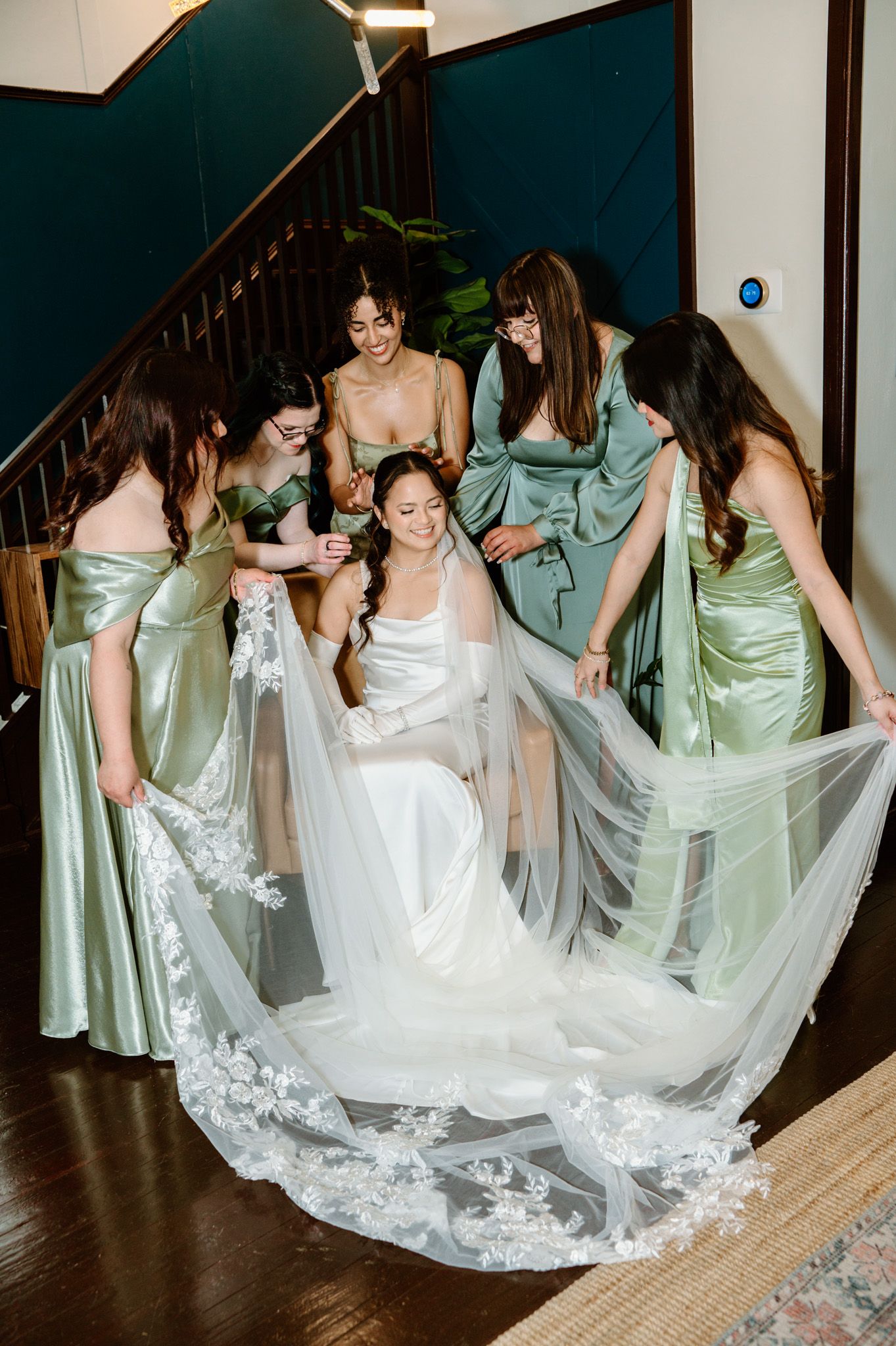 Bride seated in white gown with lace veil surrounded by bridesmaids in sage green satin dresses helping arrange her veil.