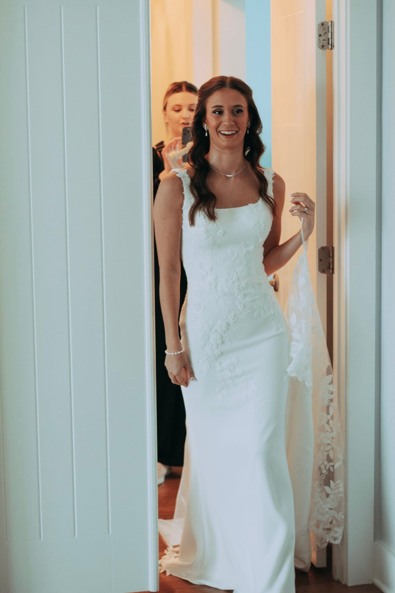 Smiling bride in elegant white lace wedding dress standing in doorway with bridesmaid behind her.