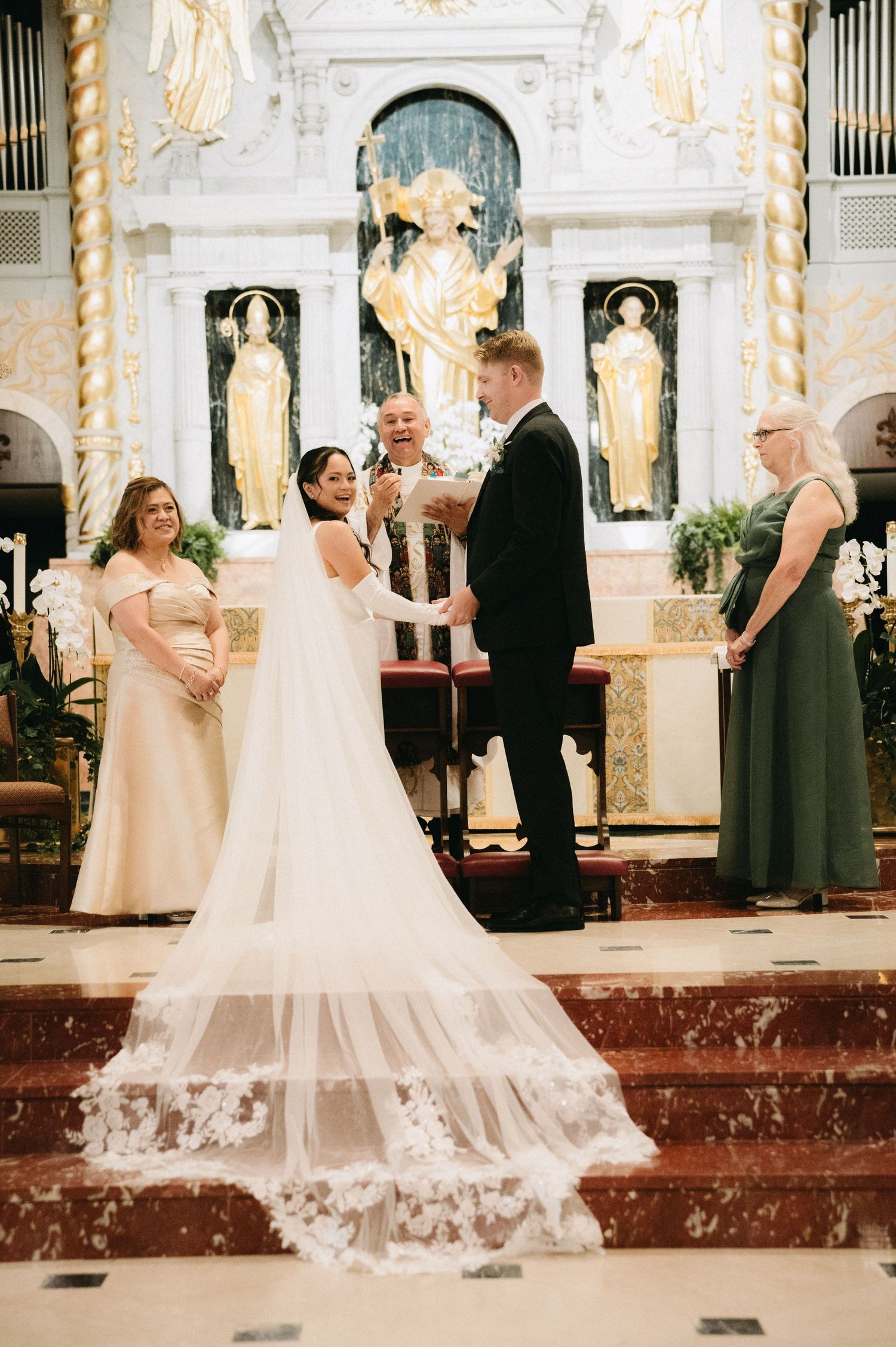 Bride and groom exchange vows at ornate church altar, bride wearing flowing gown with lace-trimmed cathedral veil.