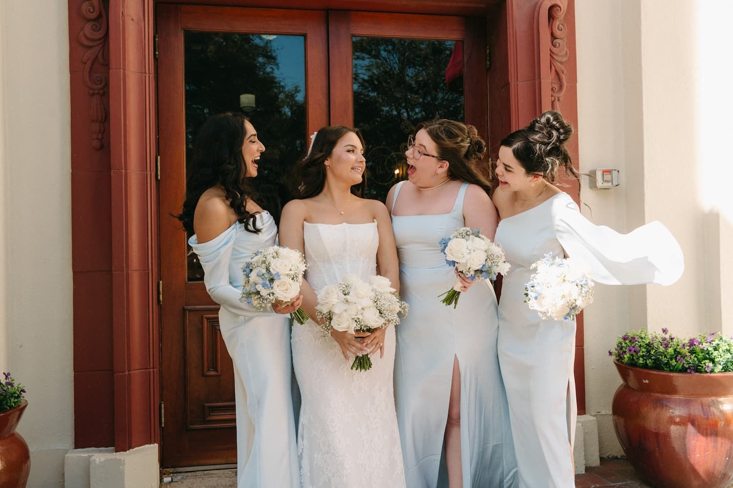 Bride in white gown with bridesmaids in light blue dresses holding white bouquets outside elegant wooden doors.