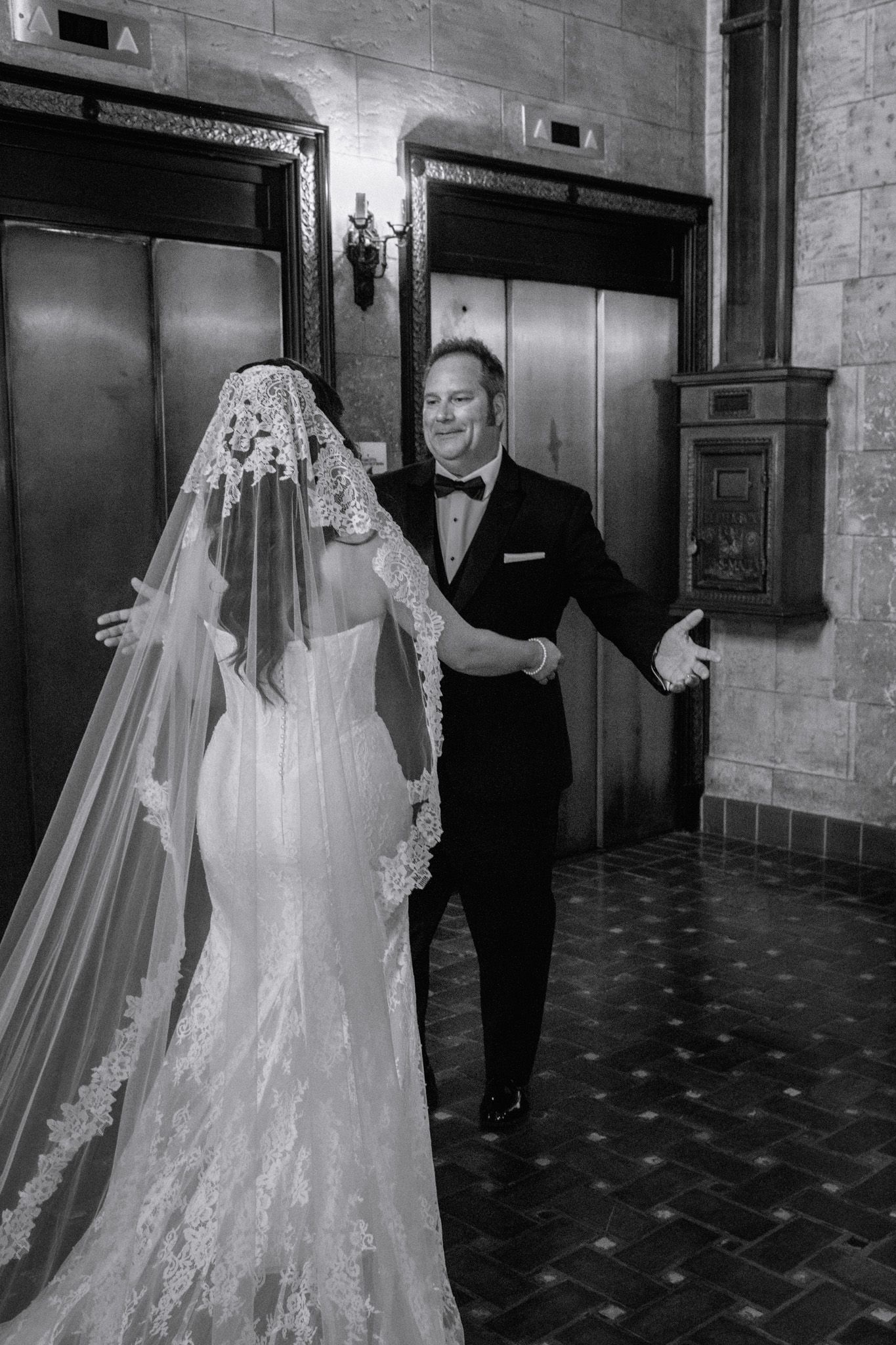 Black and white photo of a bride in lace gown and veil meeting a man in tuxedo for a first look near elevator doors.