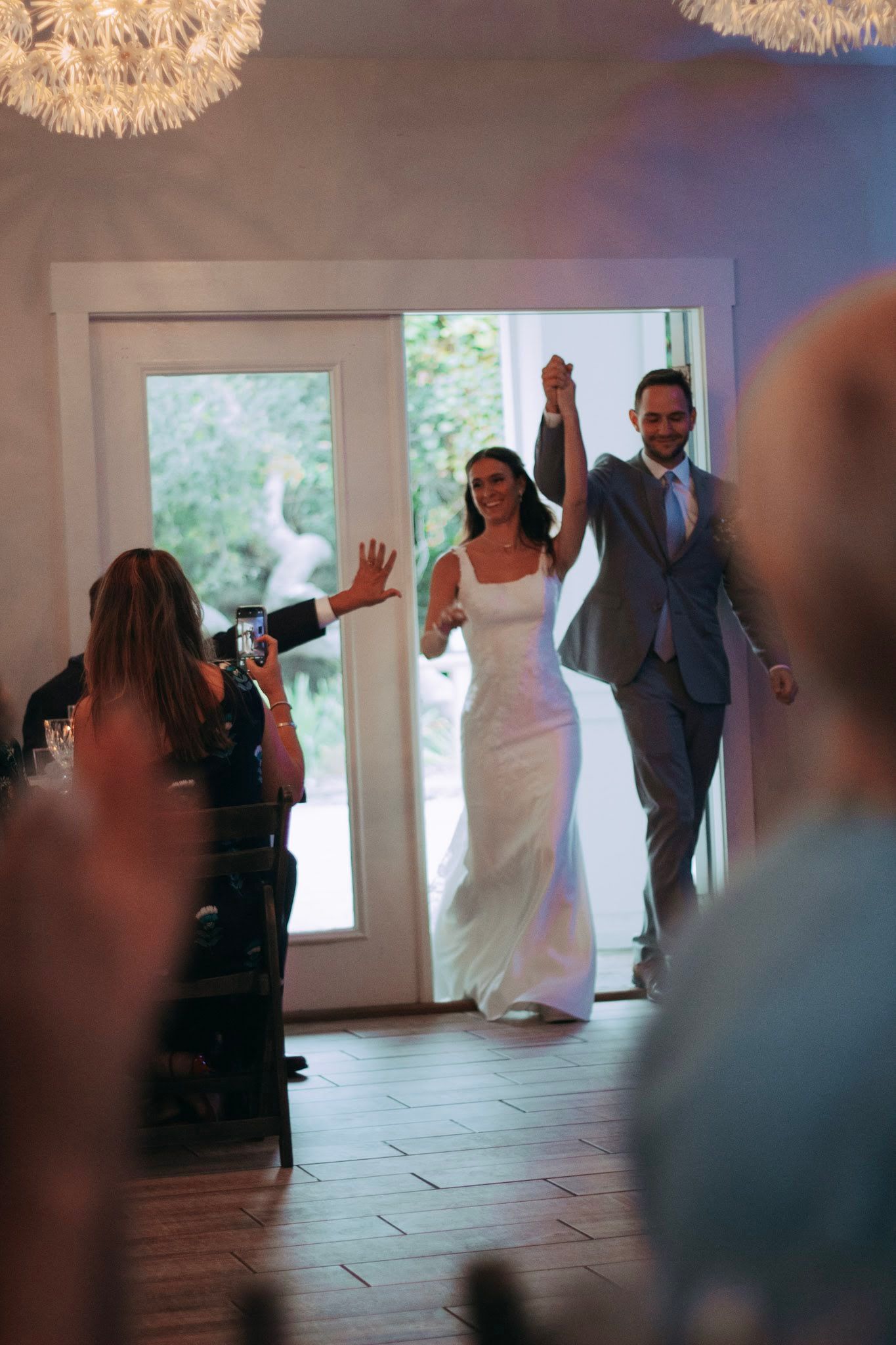 Joyful bride and groom entering reception hall with hands raised to cheering guests.
