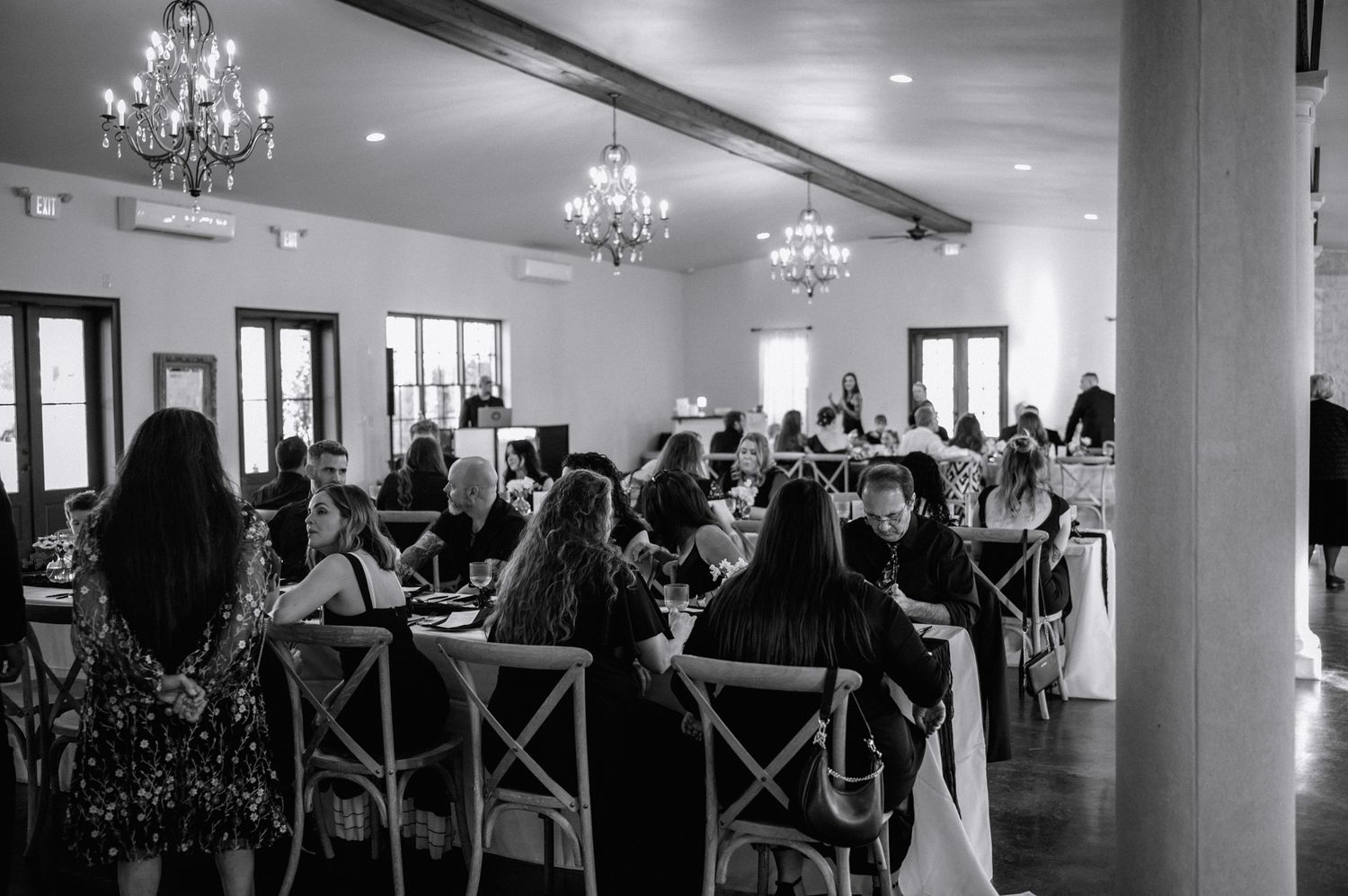 Elegant wedding reception hall with chandelier lighting and guests seated at decorated tables in black and white.