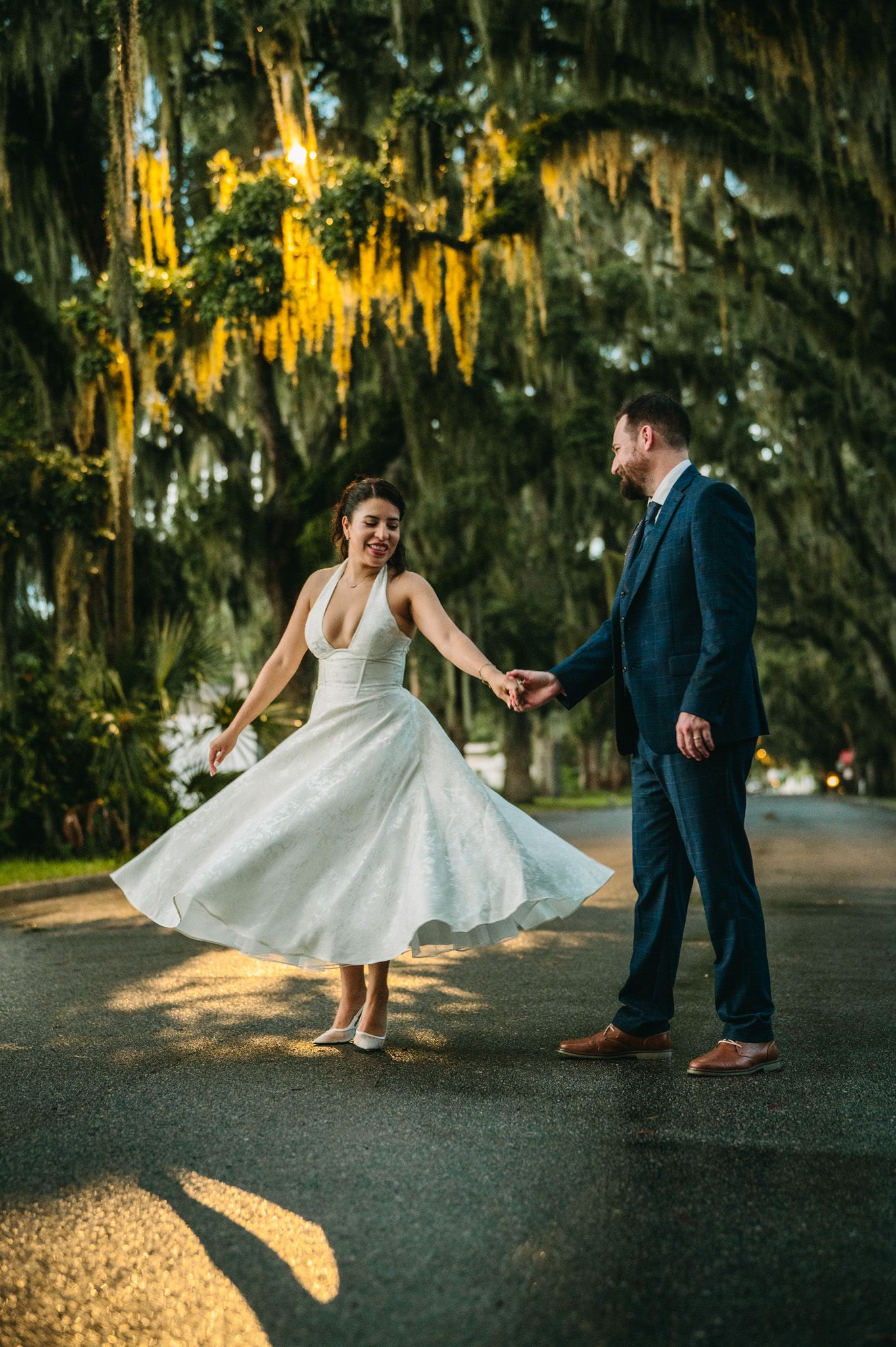 Bride twirling in tea-length dress while holding grooms hand under Spanish moss-draped trees.