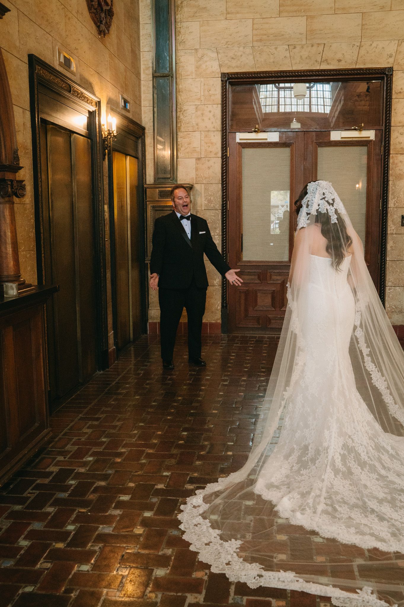 Groom reacts with joy during first look with bride in lace cathedral veil in elegant hotel corridor.