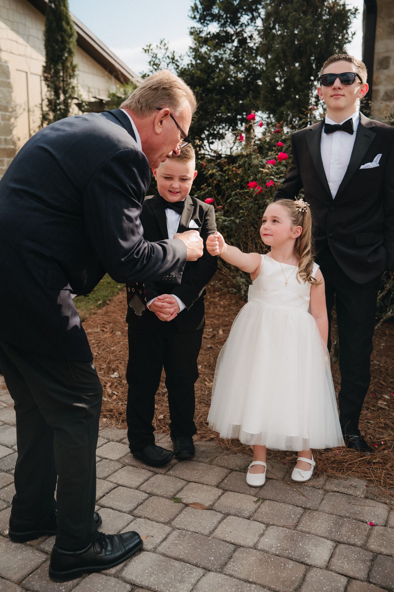 A man in a suit fist-bumps a young girl in a white flower girl dress while two boys in tuxedos stand nearby.