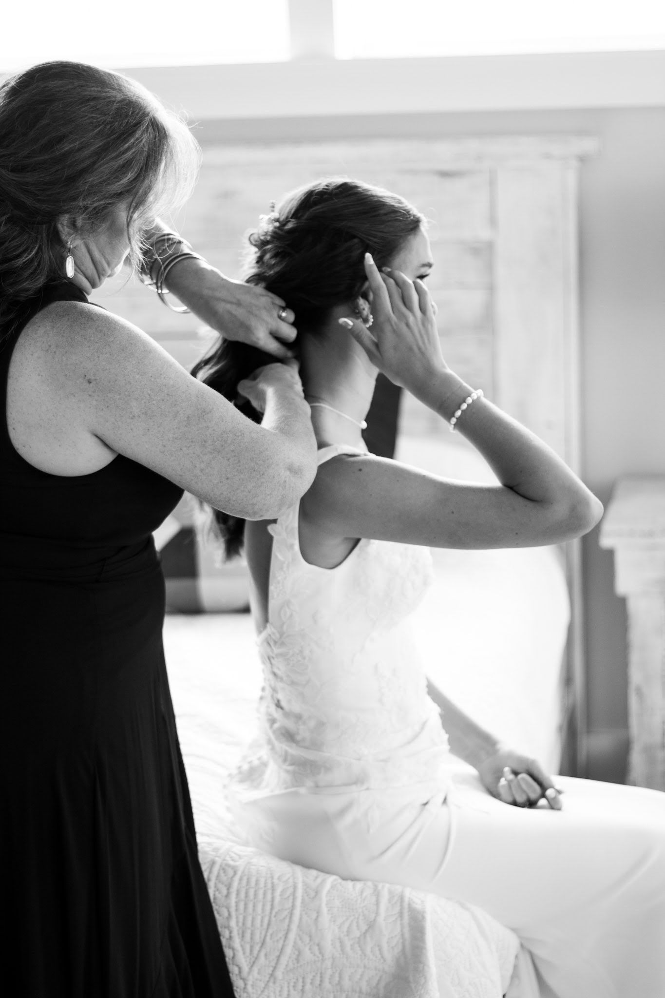 Mother helping bride with earrings while sitting on bed in black and white photo.