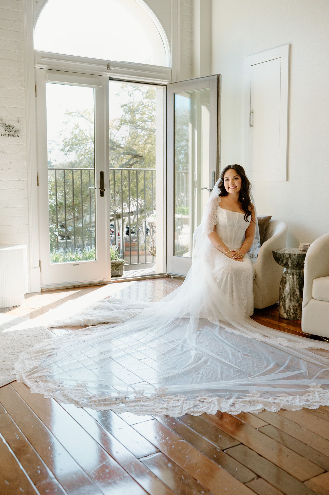 Bride in a white gown with a long lace cathedral veil spread across hardwood floors near French doors in a bright room.