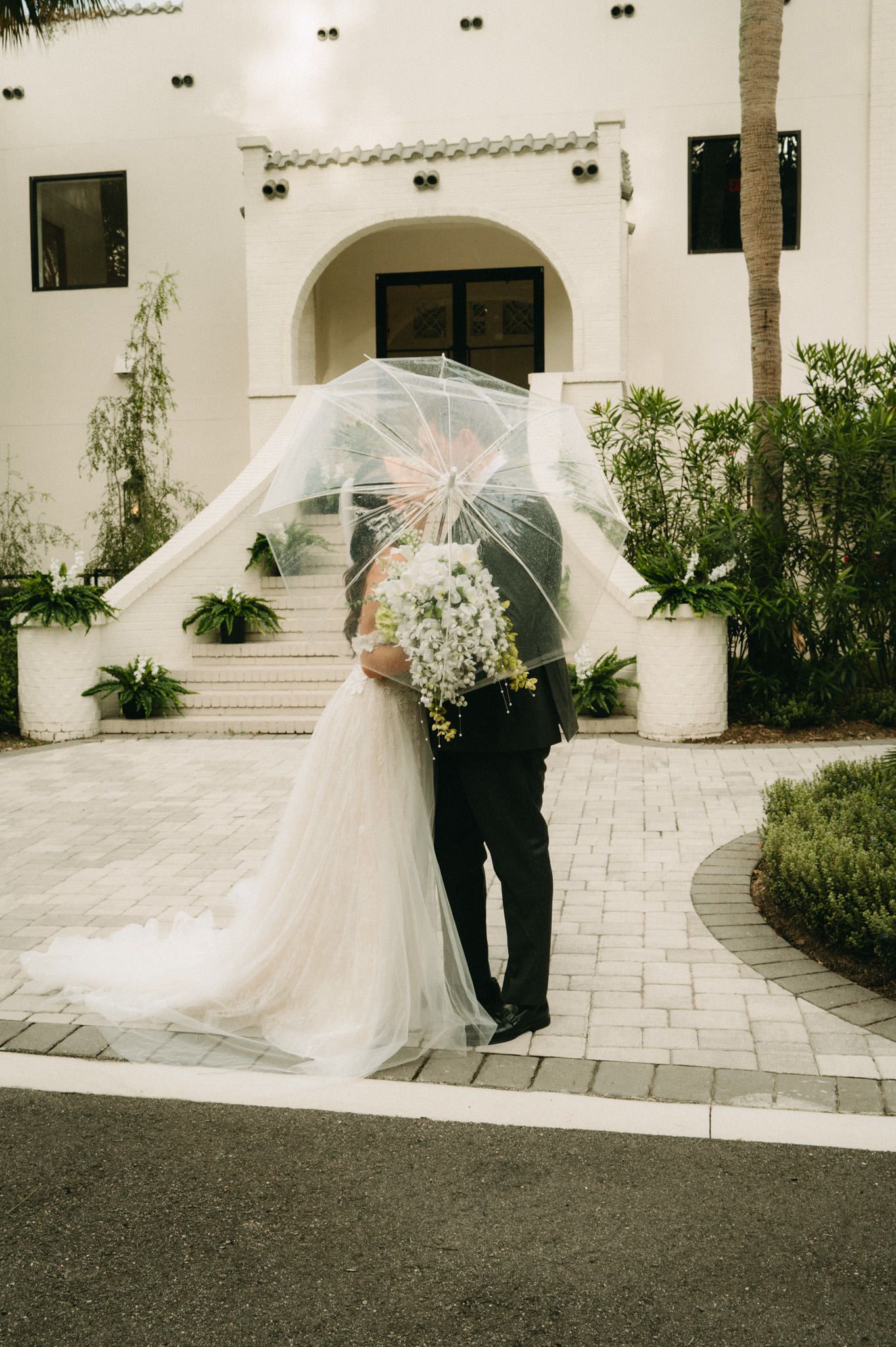 Wedding couple kisses under clear umbrella outside white Spanish-style venue.