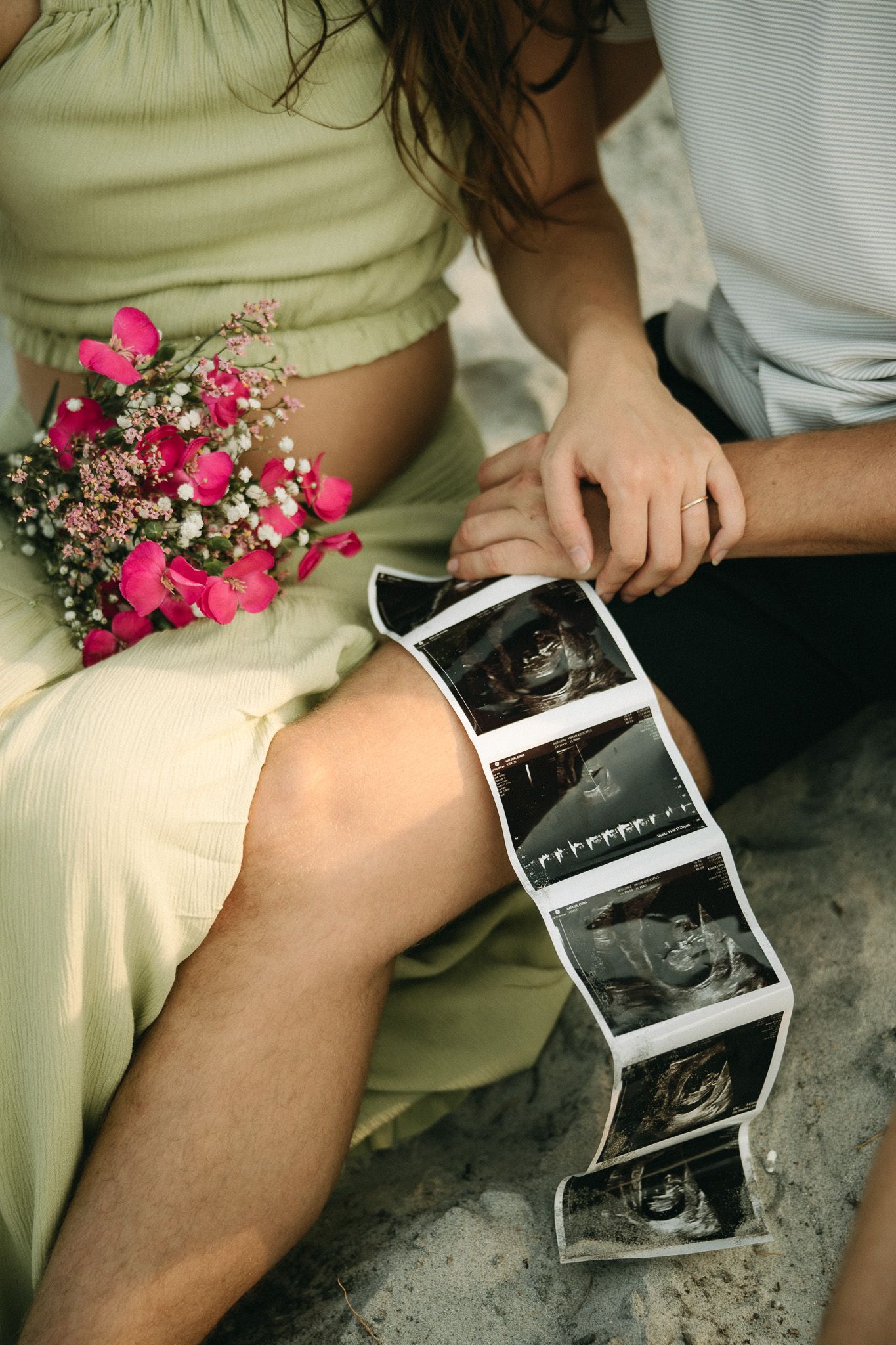 Couple holding ultrasound photos and pink flowers, celebrating pregnancy announcement while sitting together outdoors.