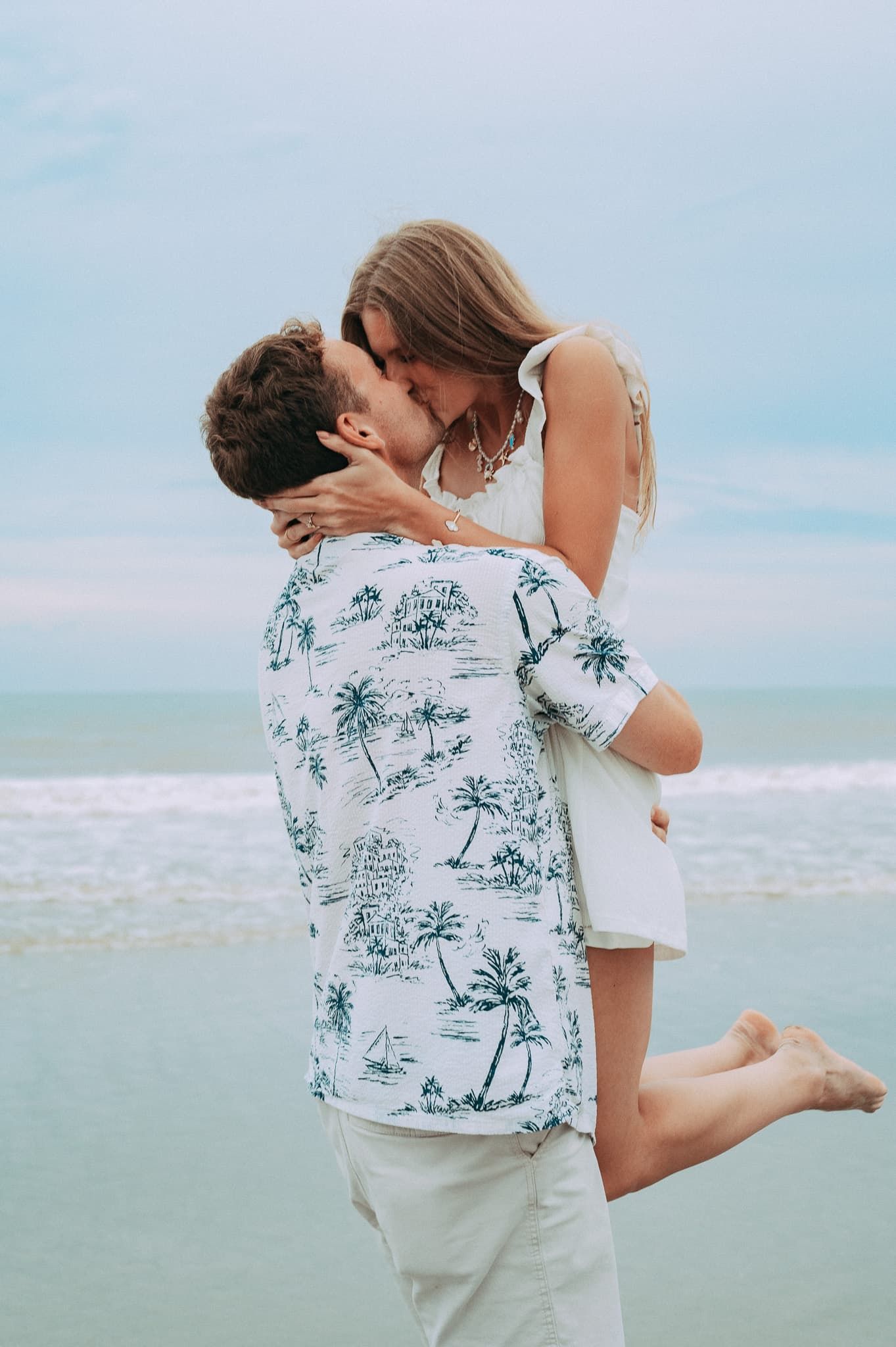 Newly engaged fiancée jumping into the arms of fiancé and share a kiss as the waves of the ocean crash behind them.