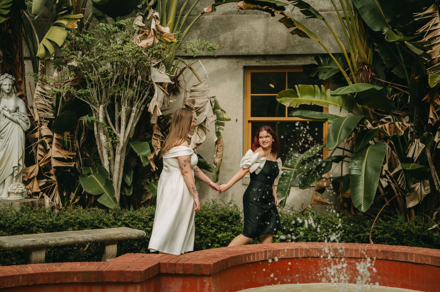 Two women in elegant dresses hold hands by a fountain surrounded by lush tropical greenery.