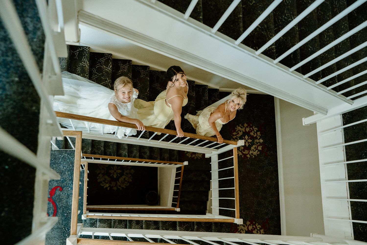 Three women in elegant dresses posing on a white spiral staircase, viewed from above in a stylish interior.