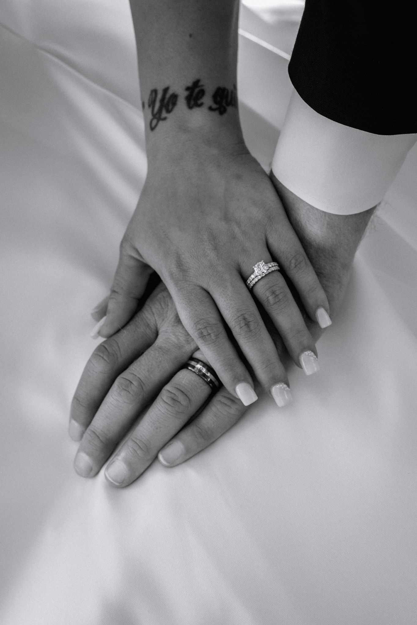 Newlyweds display wedding rings on joined hands with bride showing Yo te amo wrist tattoo in black and white.