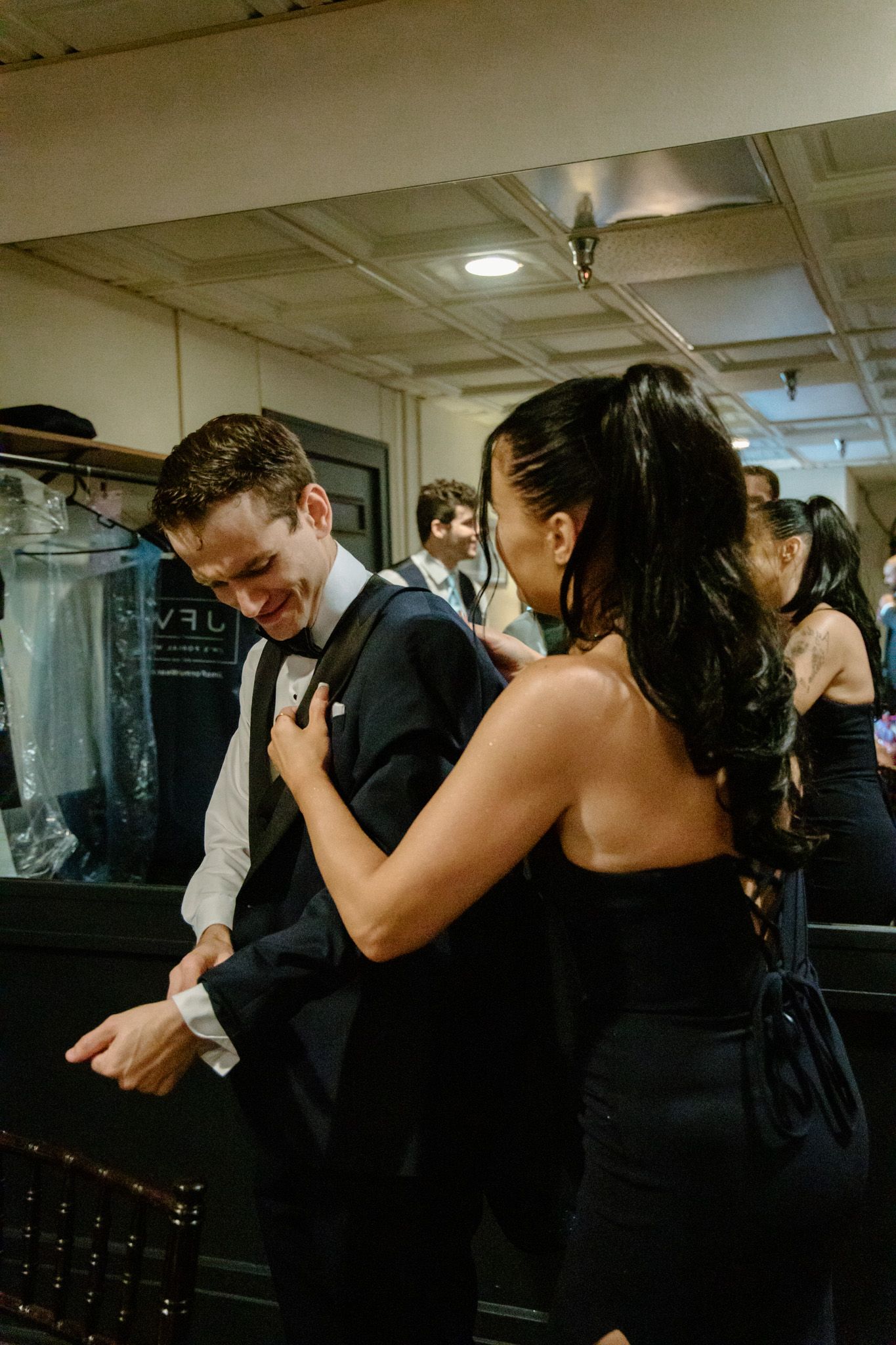 A man in a tuxedo smiles as a woman in black adjusts his bow tie backstage at a formal event.
