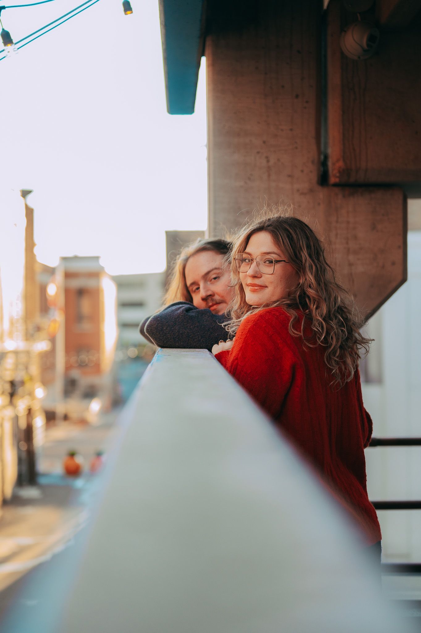 Couple leaning on a balcony railing at golden hour in an urban cityscape setting.