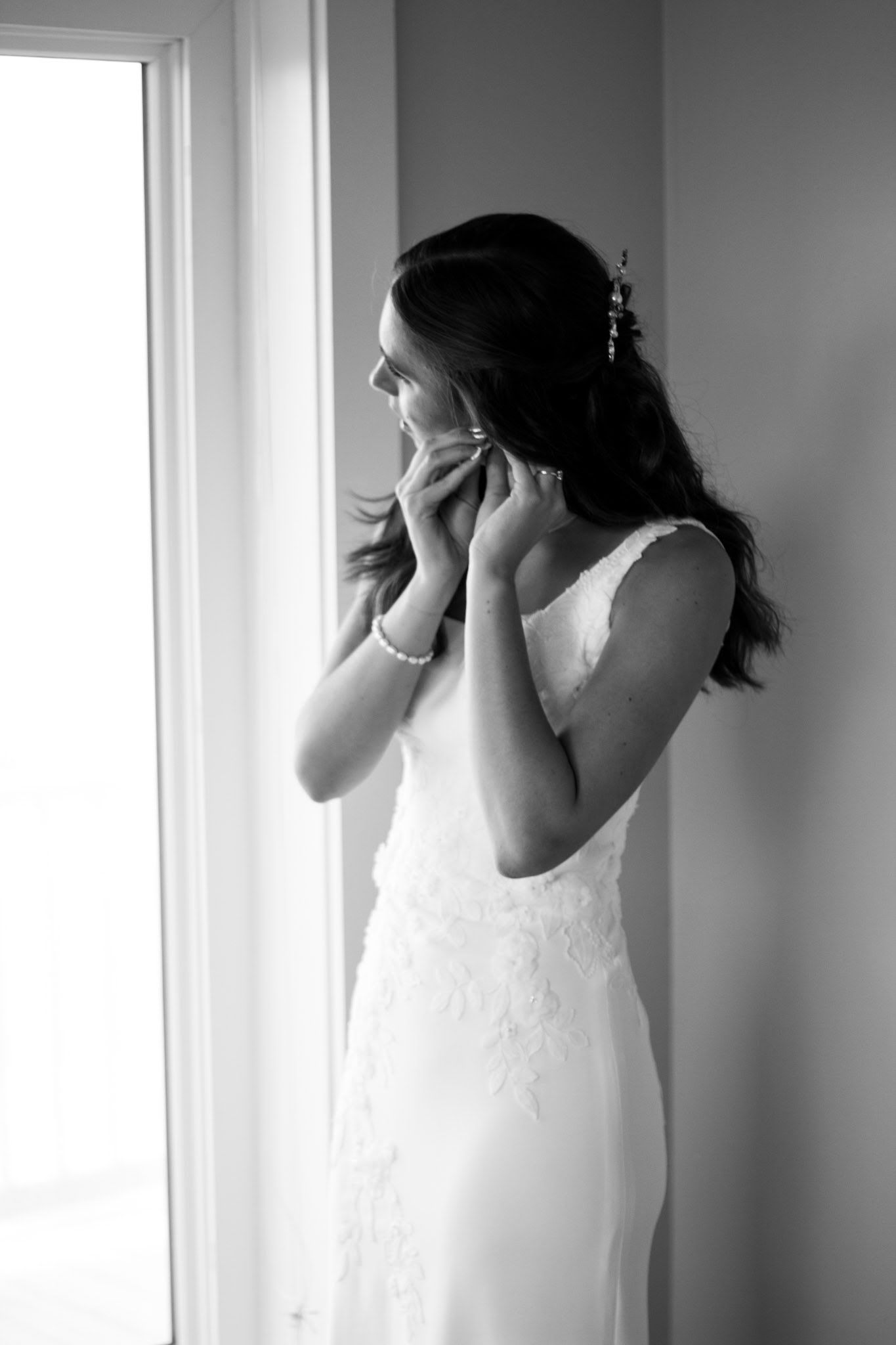 Bride in white lace gown adjusting earring by bright window in black and white photo.