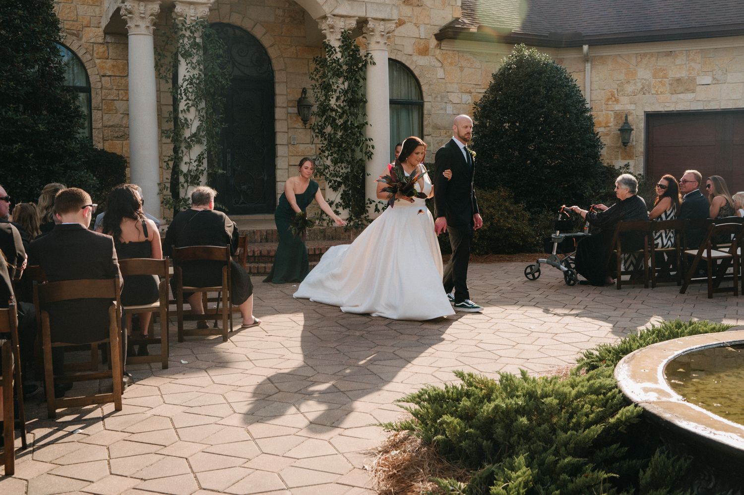 Bride walks down the aisle escorted by groom on a stone courtyard patio at an outdoor wedding ceremony.