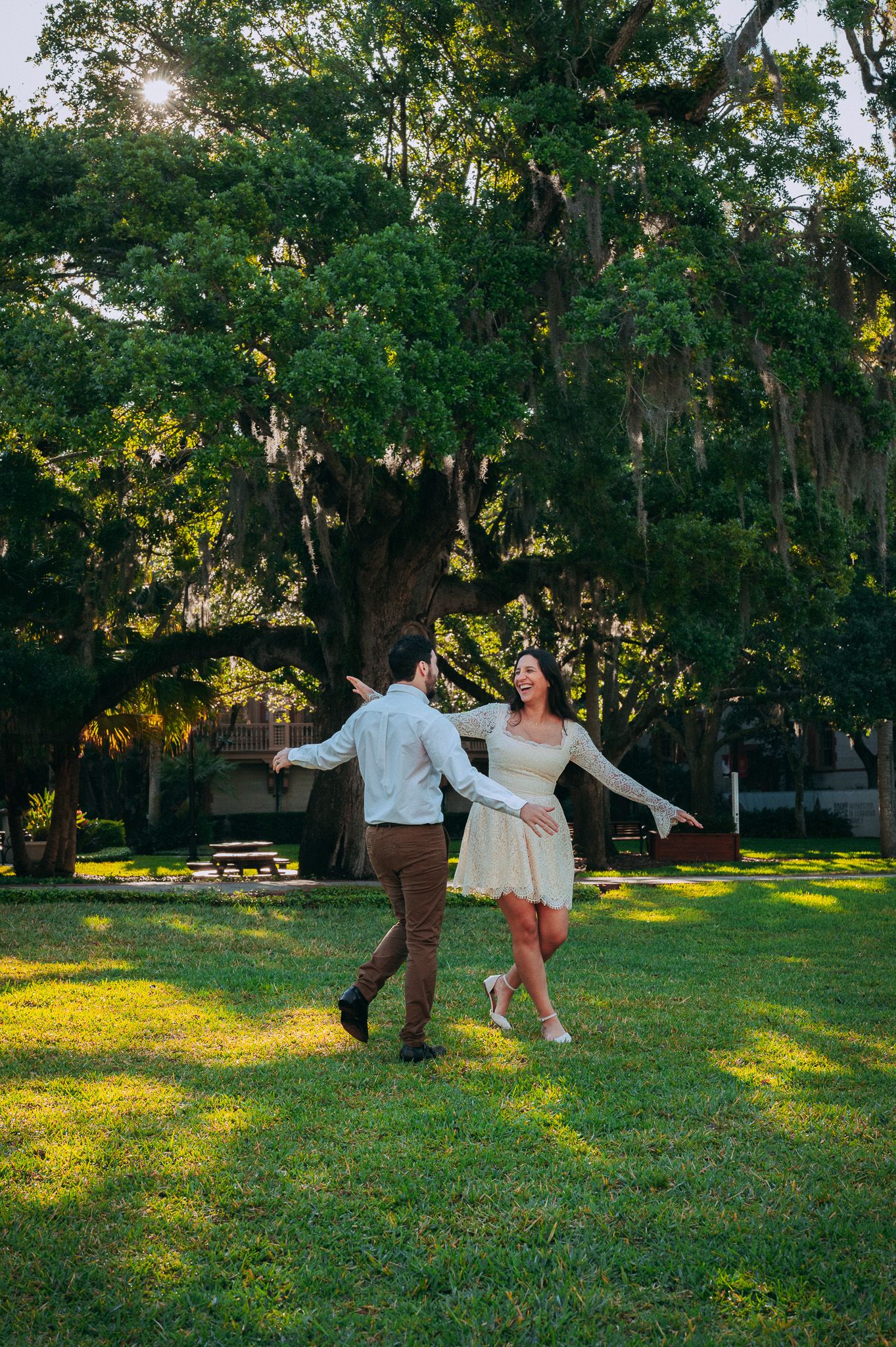 Couple joyfully dancing on a sunlit green lawn surrounded by large moss-draped trees.