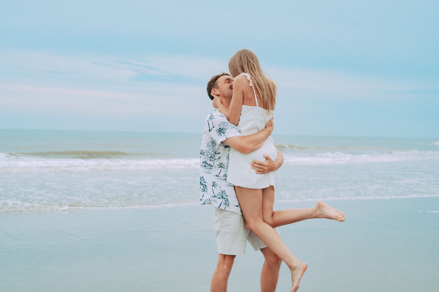 Romantic couple embracing and kissing on a serene beach with ocean waves in the background.