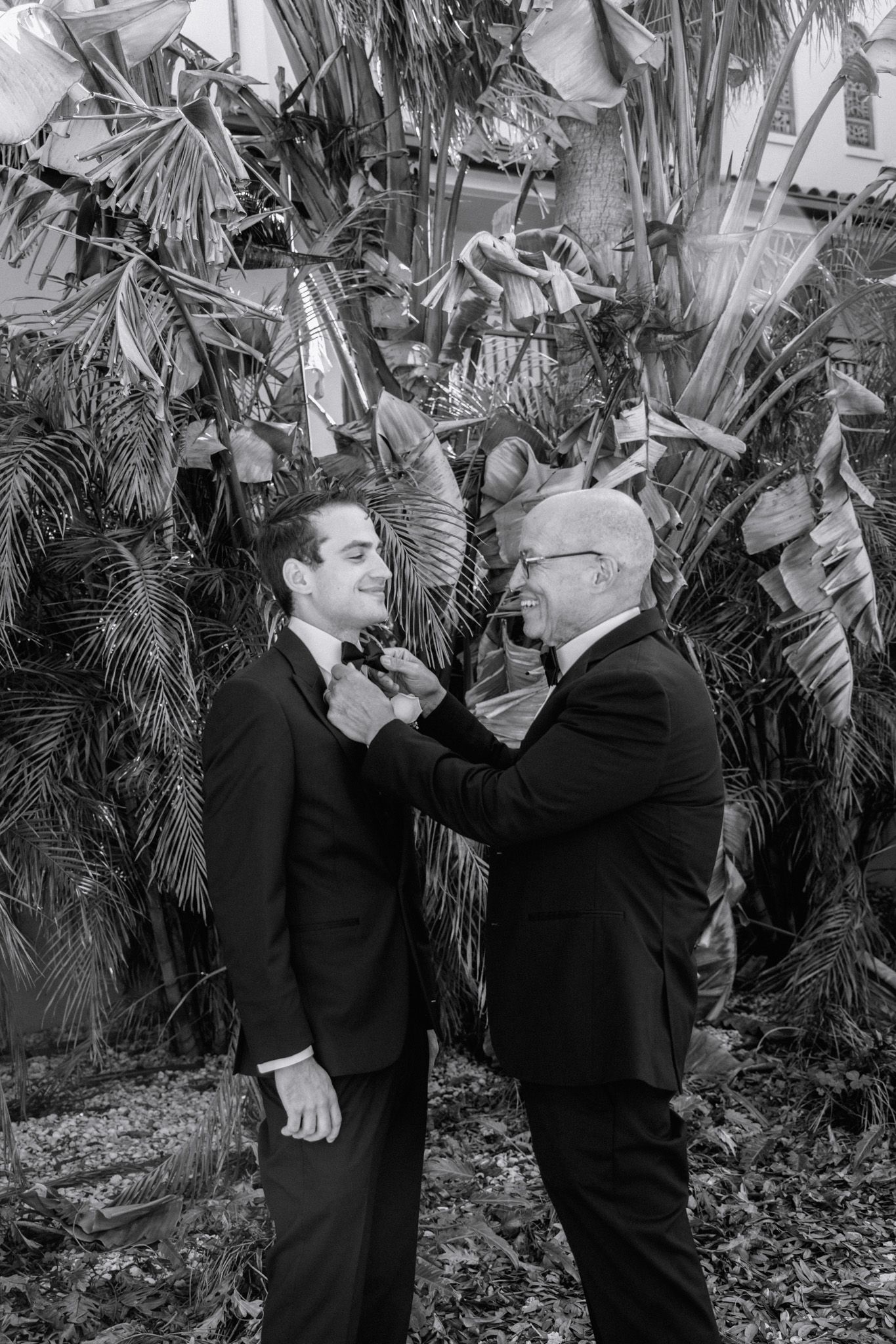 Black and white photo of two men in tuxedos, one adjusting the other's bow tie outdoors near tropical plants.