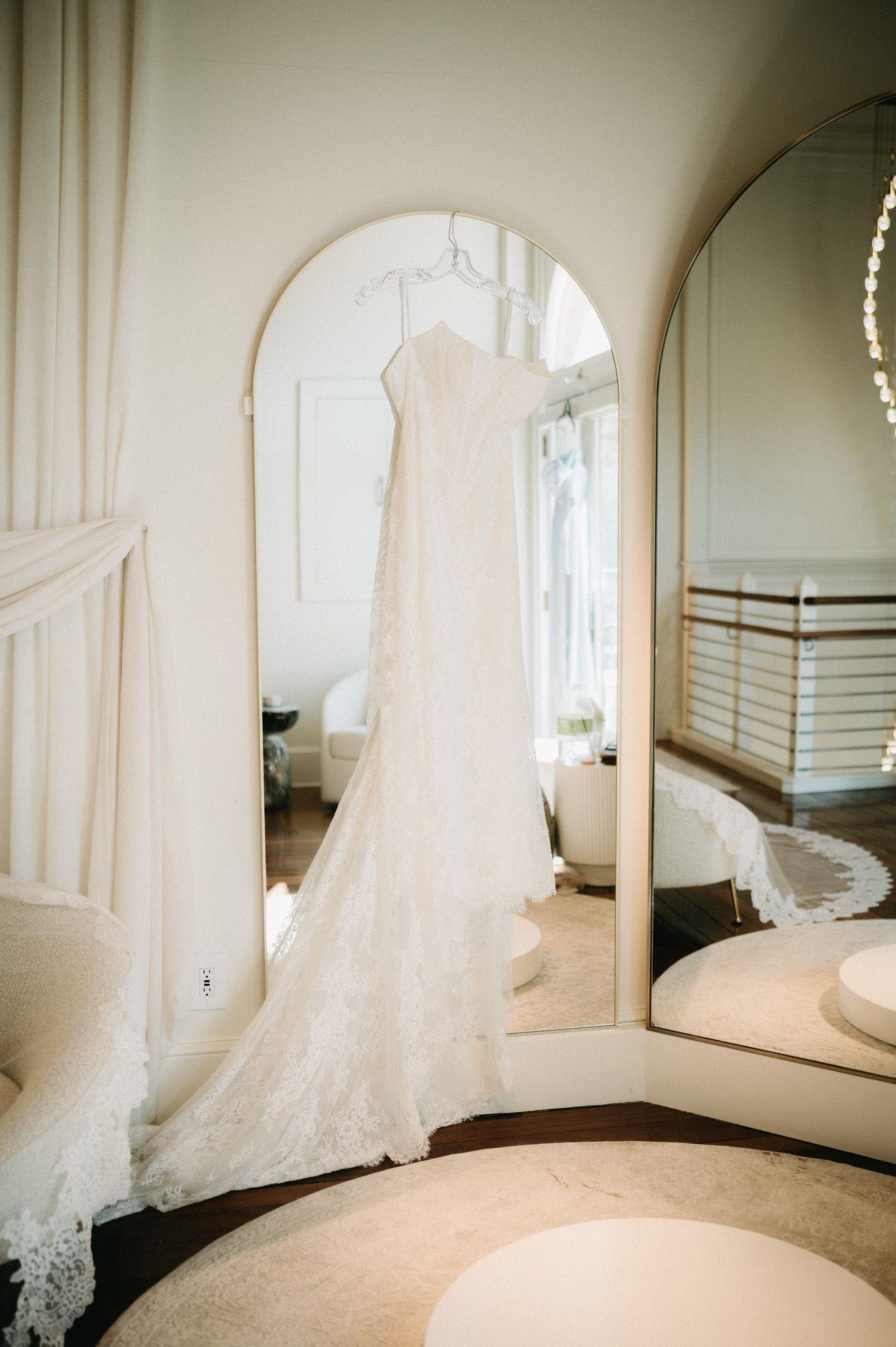 Elegant white lace wedding dress hanging in an arched mirror in a bright bridal suite.