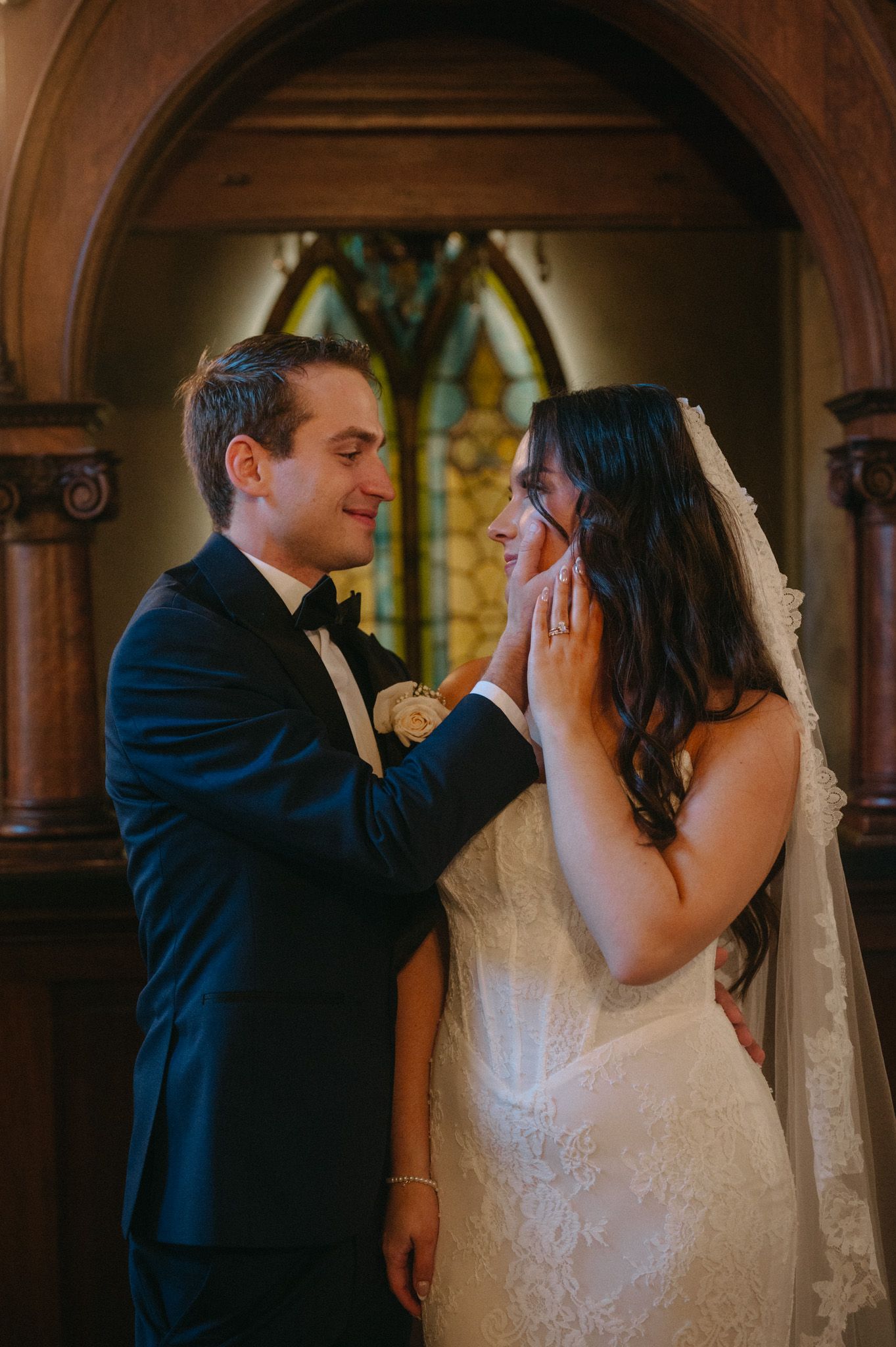 Groom tenderly cups bride's face in a church, both smiling, with stained glass and arched columns in the background.