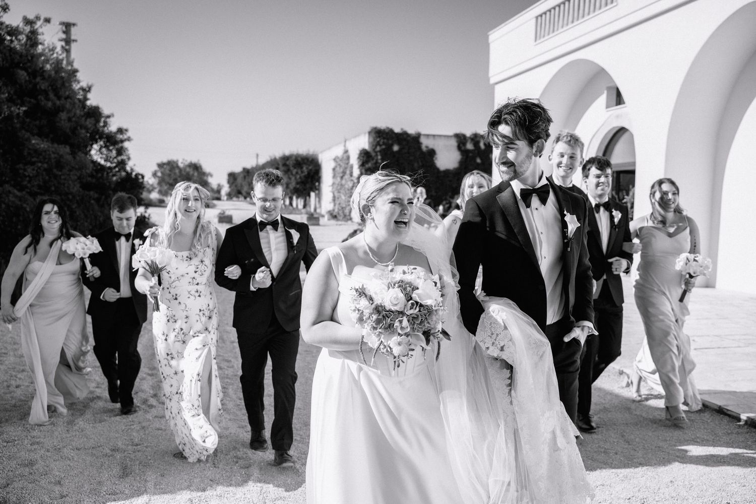 Black and white photo of a joyful wedding party walking outdoors near an arched building on a sunny day.