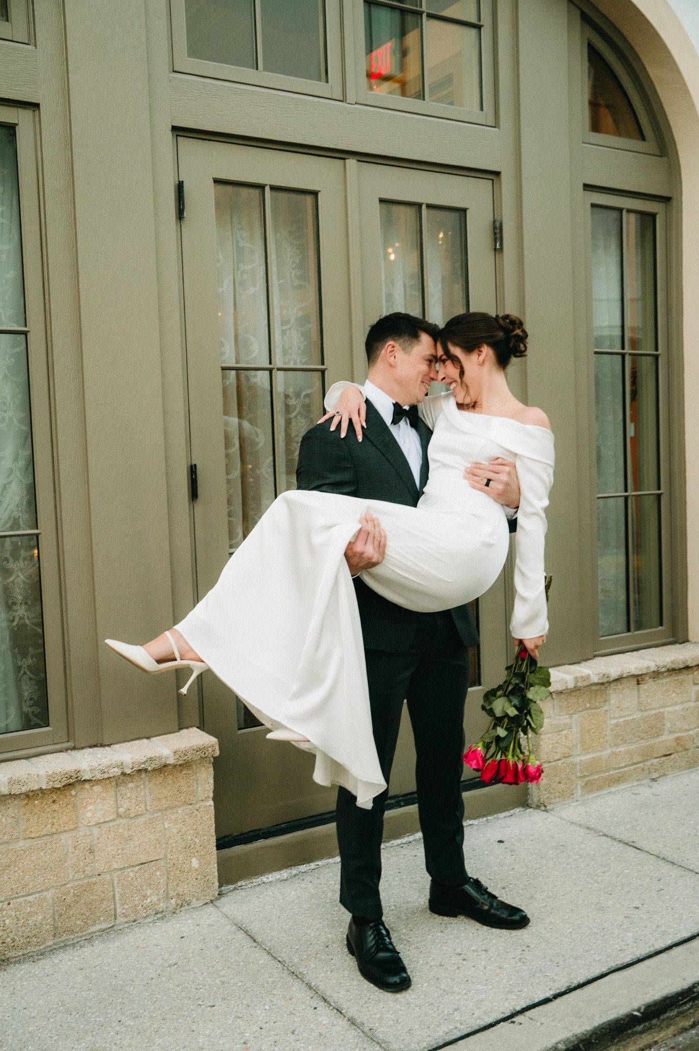Groom carries bride in white off-shoulder gown holding pink roses outside elegant building.