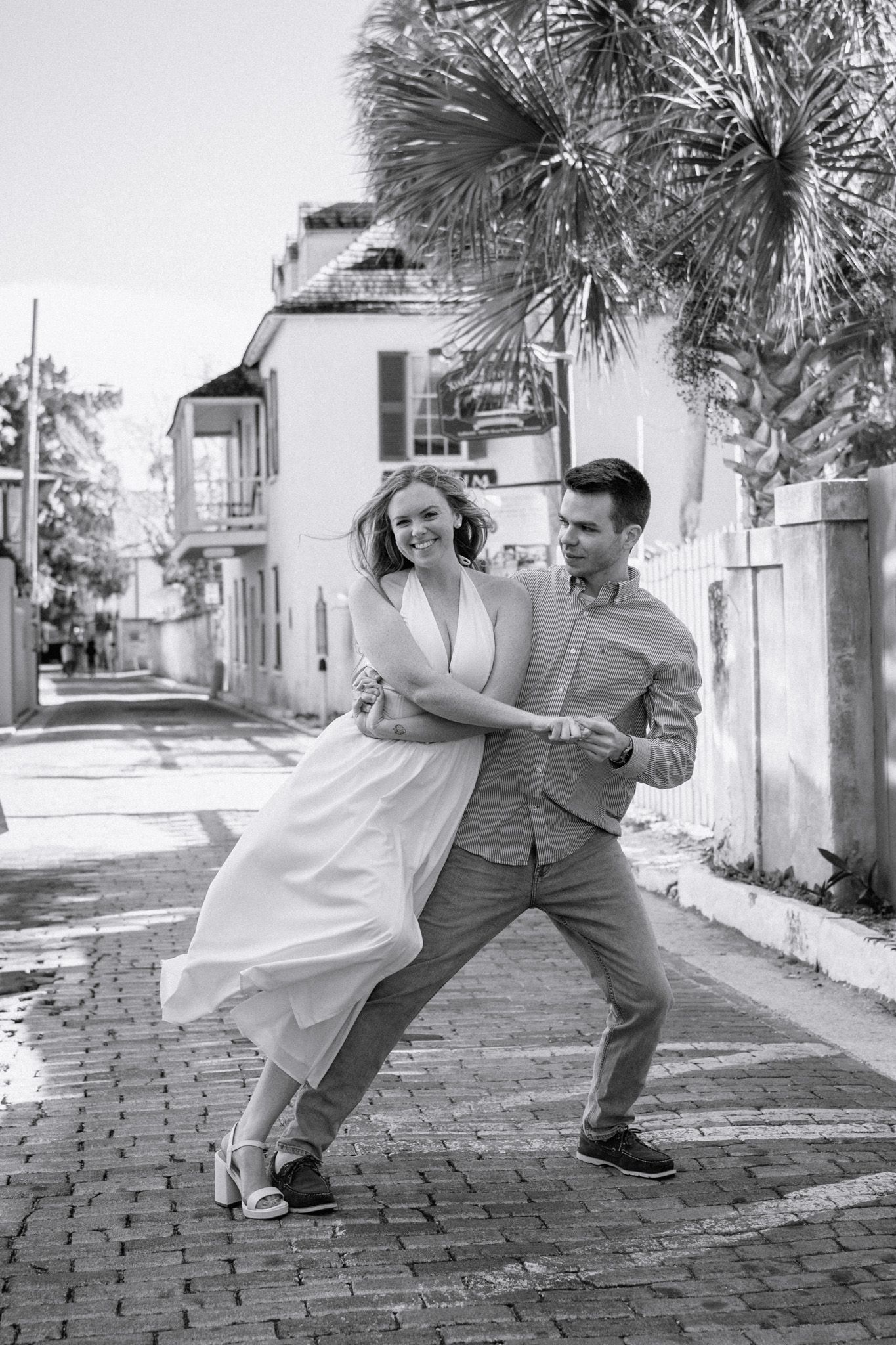 Black and white photo of a couple dancing joyfully on a cobblestone street with palm trees and historic buildings behind them.