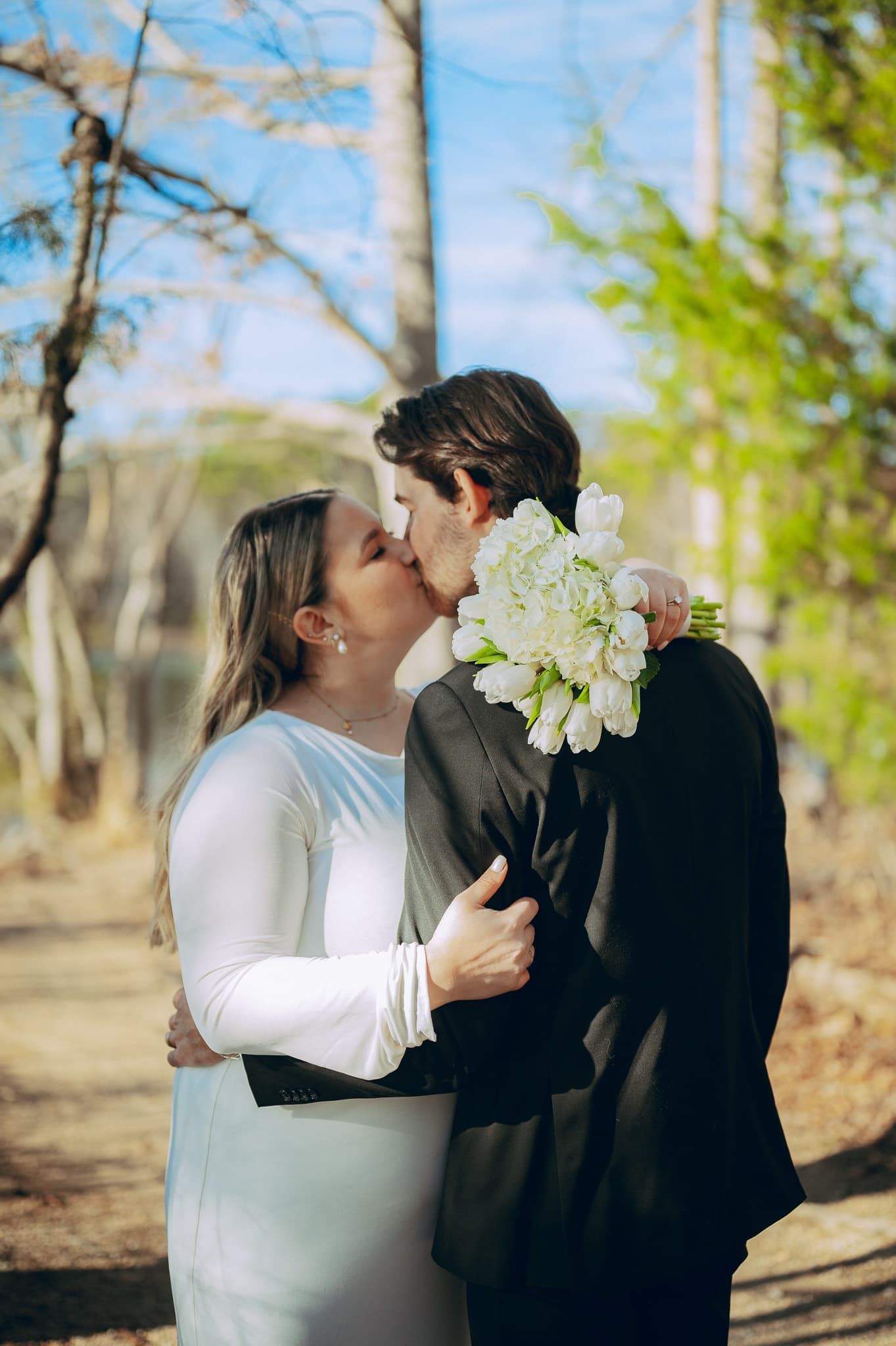 Bride and groom sharing a tender kiss outdoors, bride holding white tulip bouquet in woods.