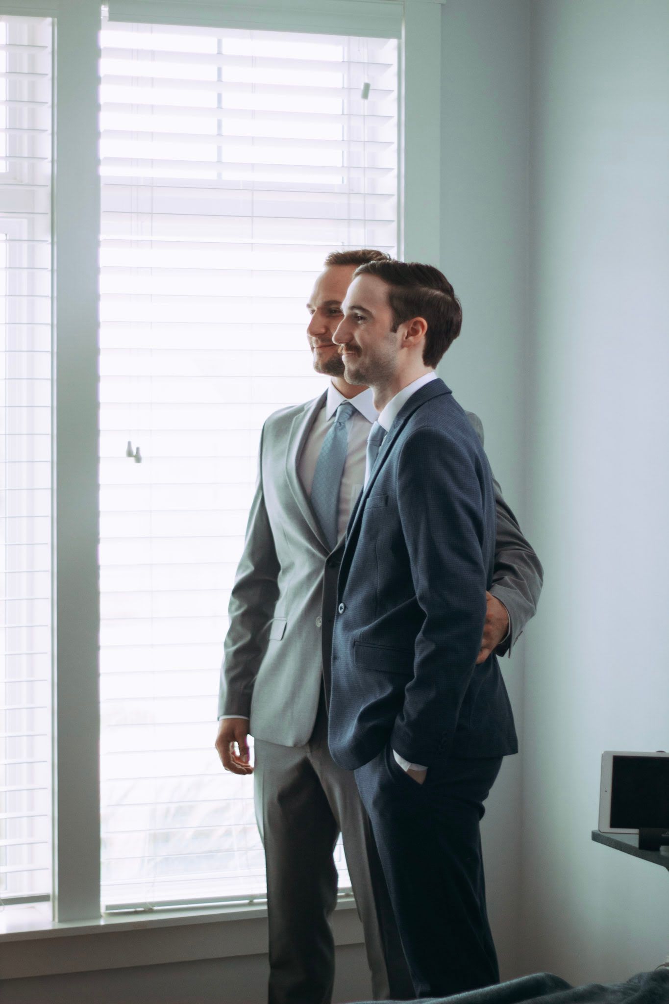 Two groomsmen in gray and navy suits standing together looking out bright window.
