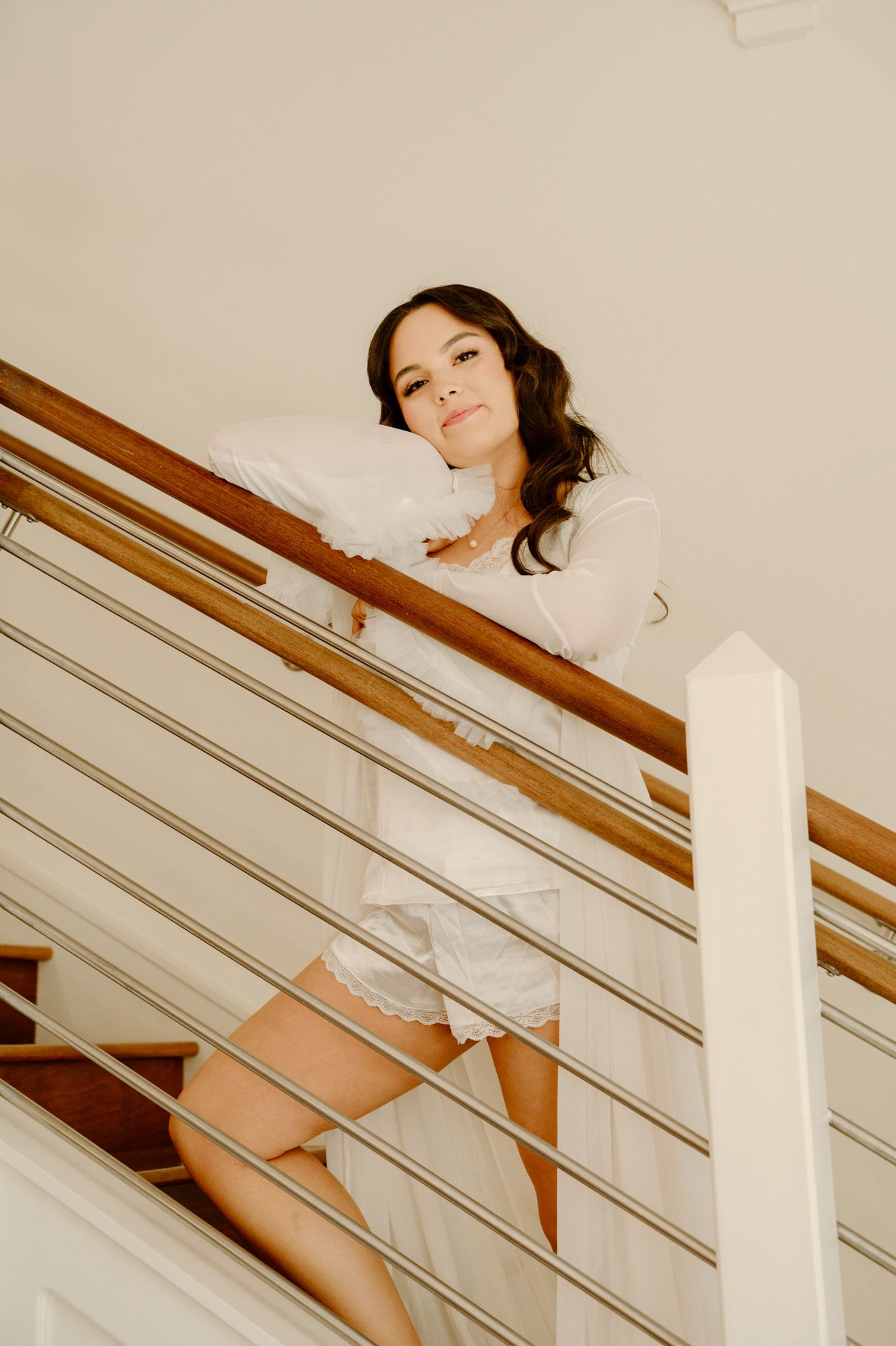 A woman in a white dress leans on wooden stair railing, photographed from below in a bright interior.
