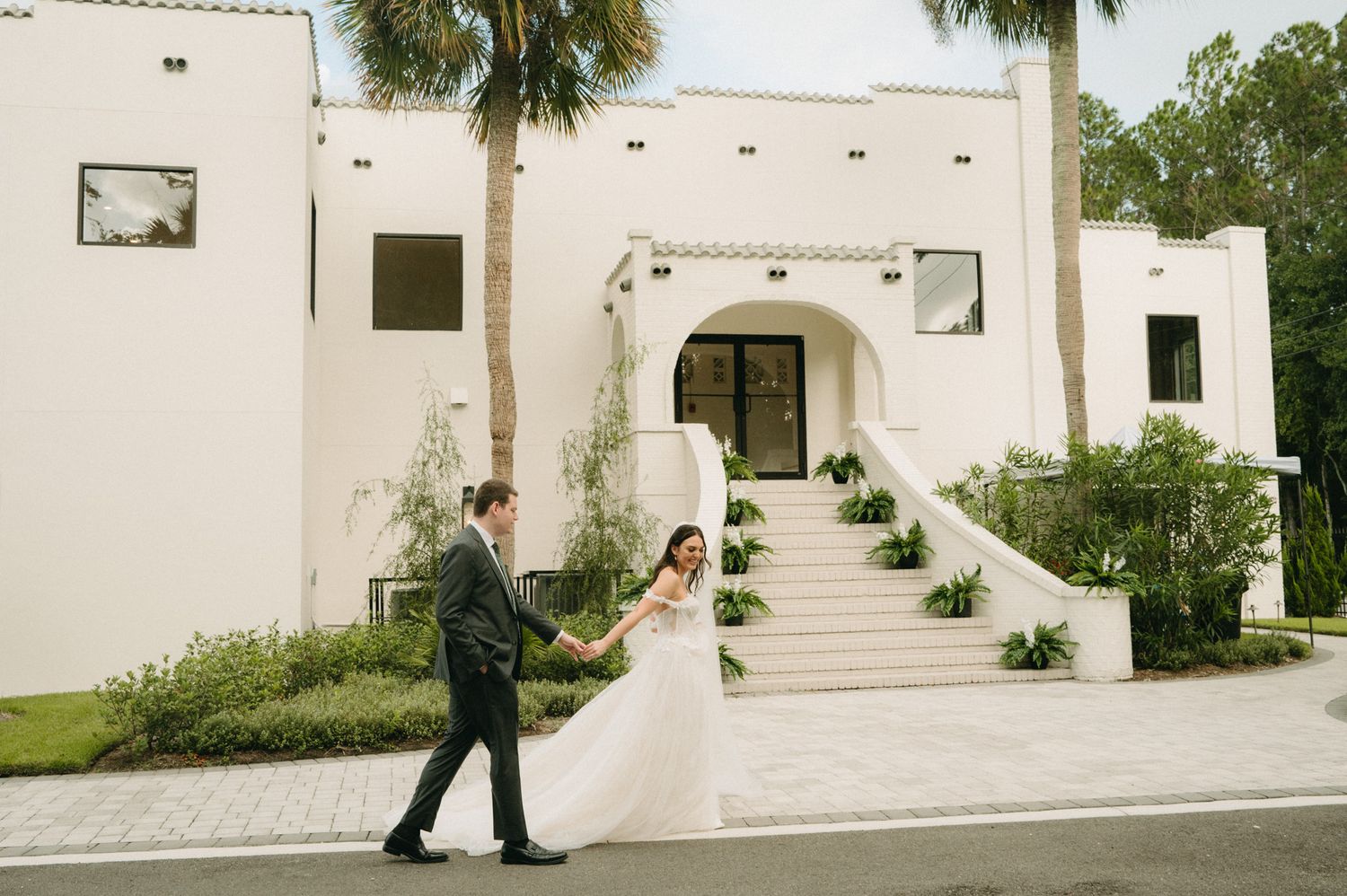 Bride in flowing white gown and groom walk hand in hand outside a white Spanish-style estate.