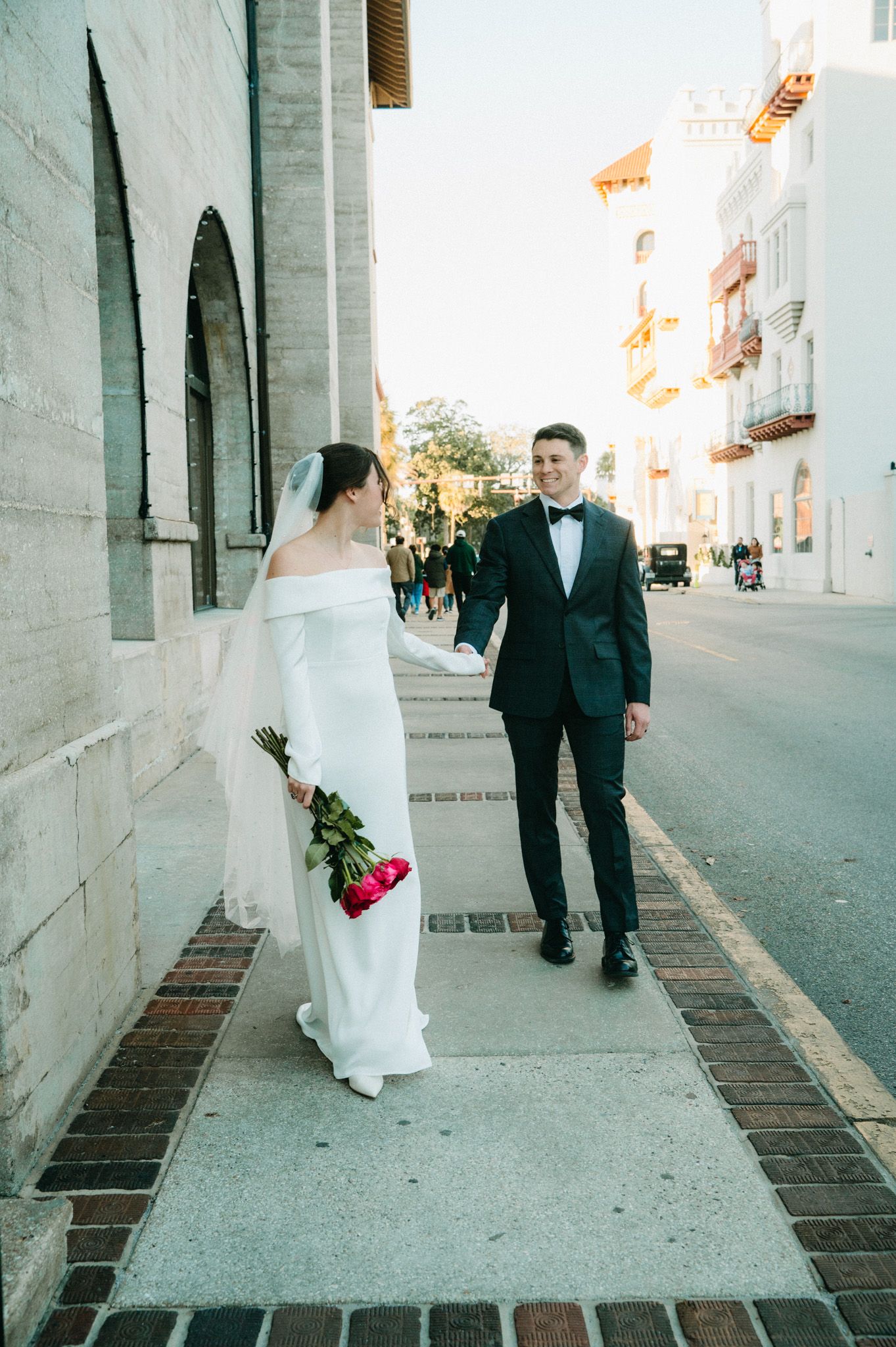 Bride in white off-shoulder gown holds pink roses while walking with tuxedo groom on sunny street.