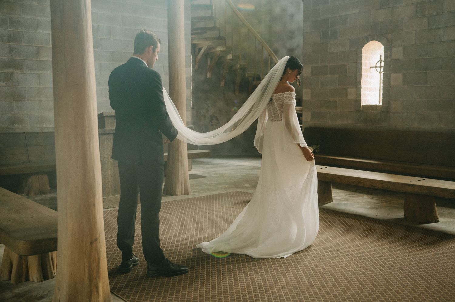 Groom gently holding his bride's long veil inside a rustic stone chapel with wooden beams.