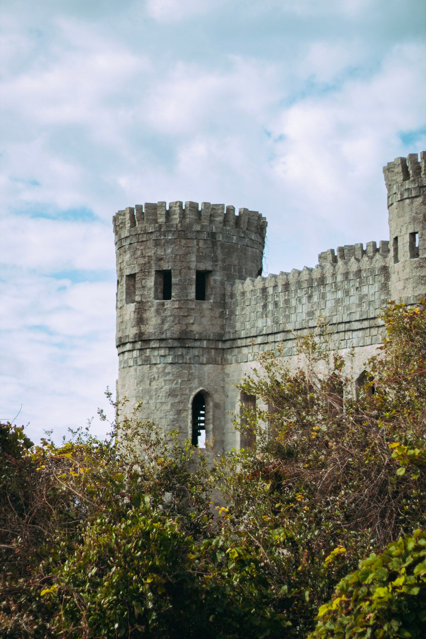 Ancient stone castle tower with battlements rising above lush green trees against cloudy sky.