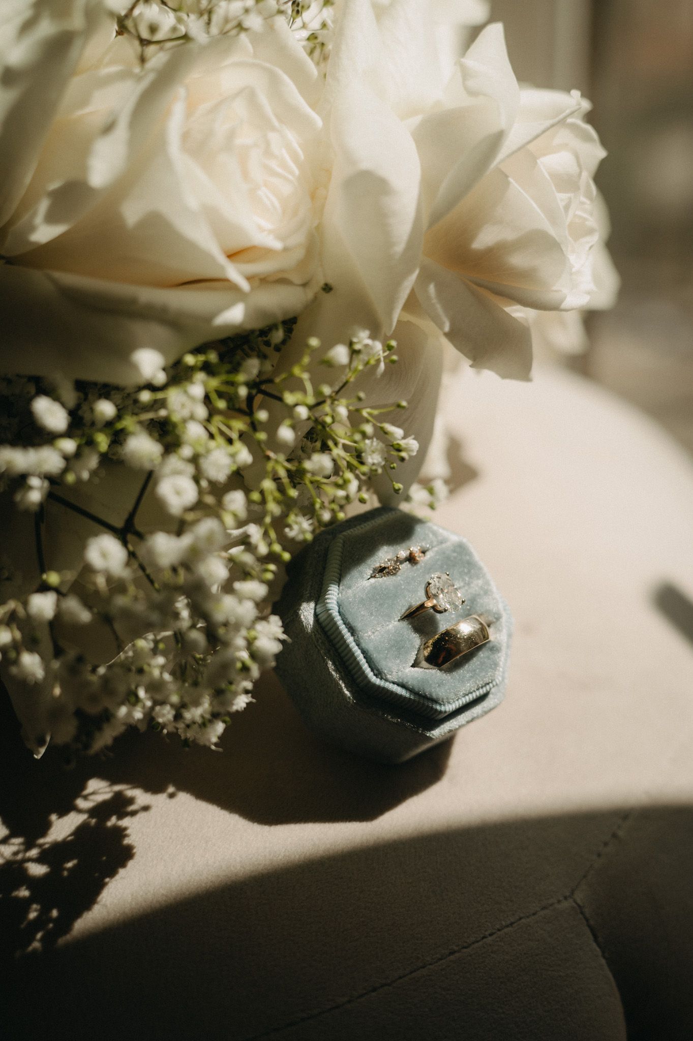 White roses and baby's breath bouquet beside a blue velvet ring box with wedding bands on a neutral surface.