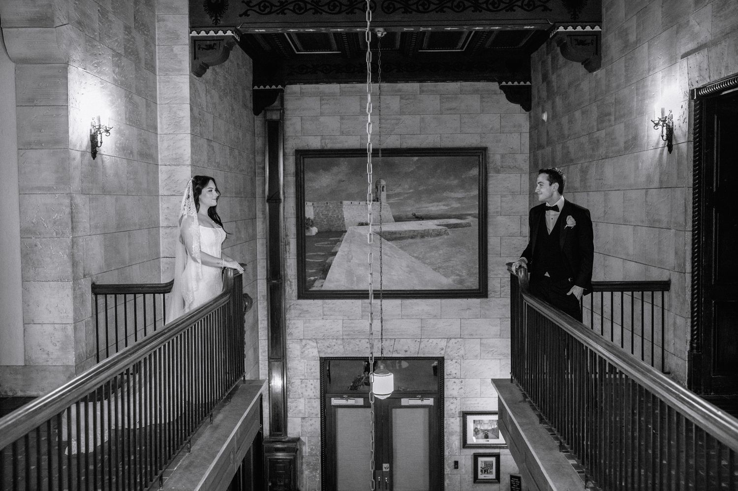 Bride and groom share a first look moment on separate staircases in an elegant stone venue in black and white.