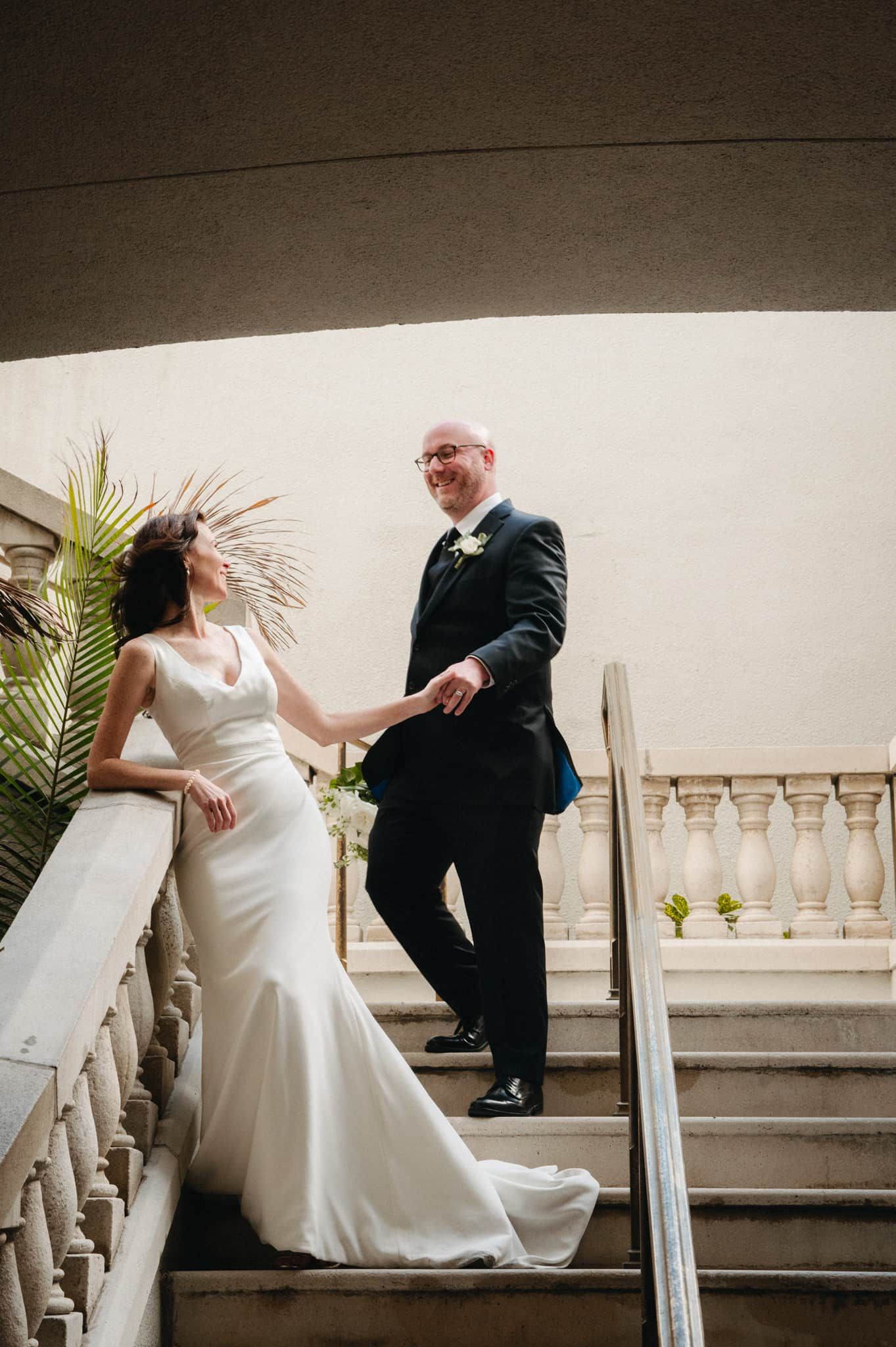 Bride and groom hold hands on elegant stone staircase, she in fitted white gown, he in dark suit, smiling joyfully at the Lightner Museum.