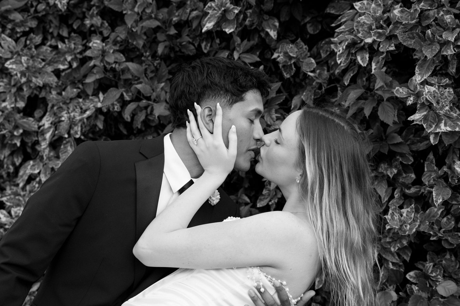 Black and white photo of a couple sharing a romantic kiss in front of lush green ivy at their wedding.