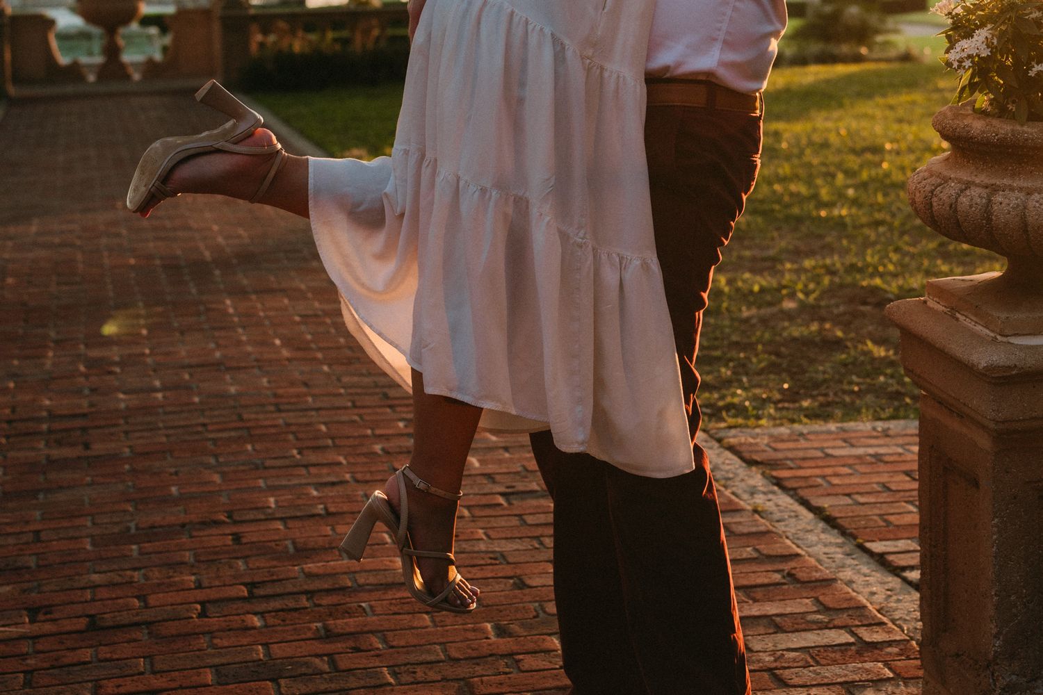 Couple embracing at golden hour on brick path, woman in white dress lifted off ground wearing silver heels.