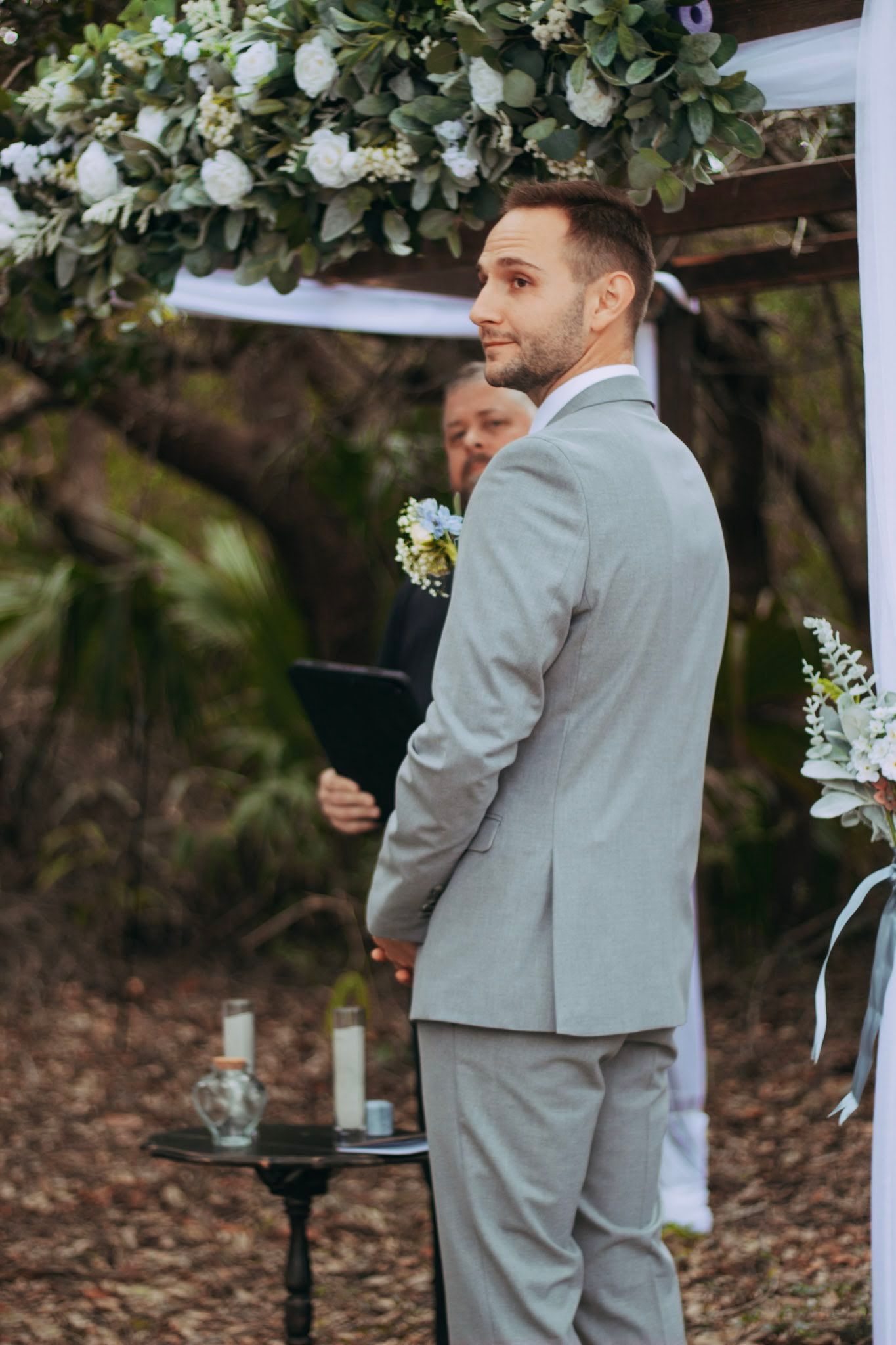 Groom in gray suit looks back smiling at wedding altar decorated with white flowers and greenery.