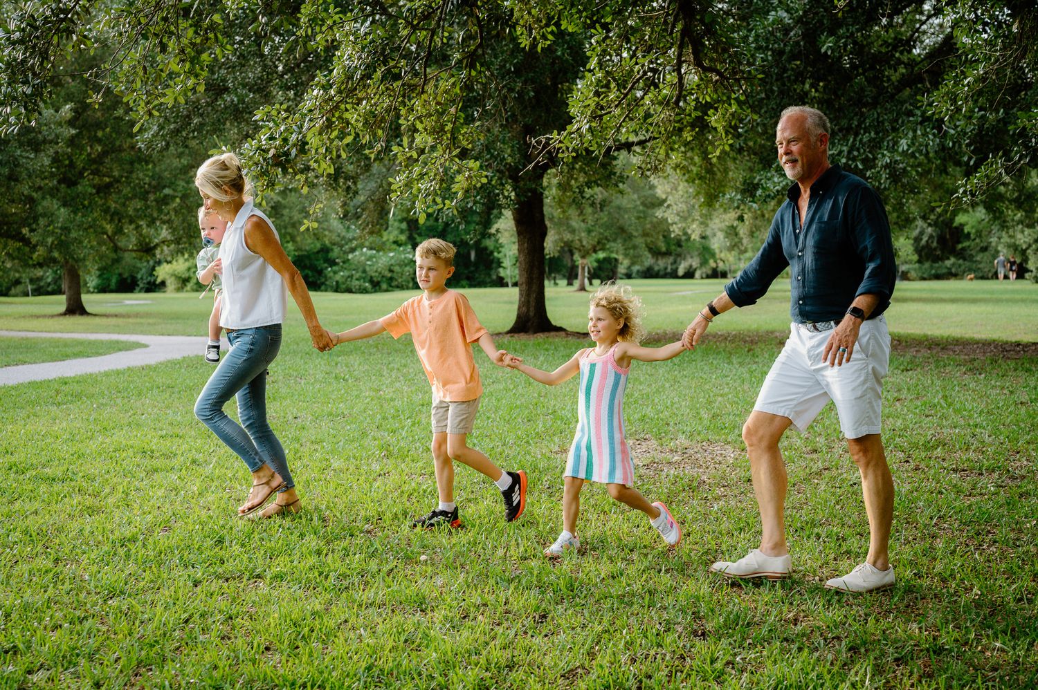 Happy family of four holding hands and walking together on green grass in a sunny park.