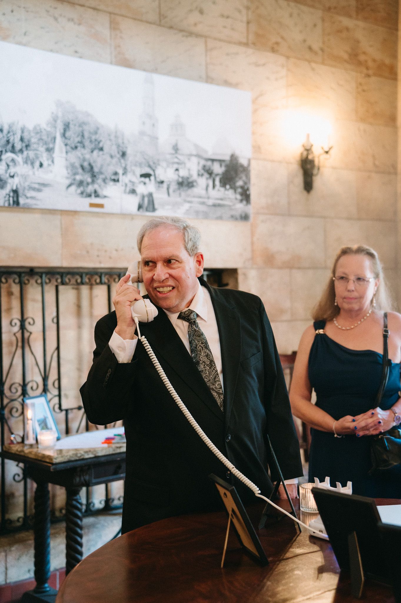 Man in tuxedo smiles while holding vintage telephone at wedding reception check-in desk.