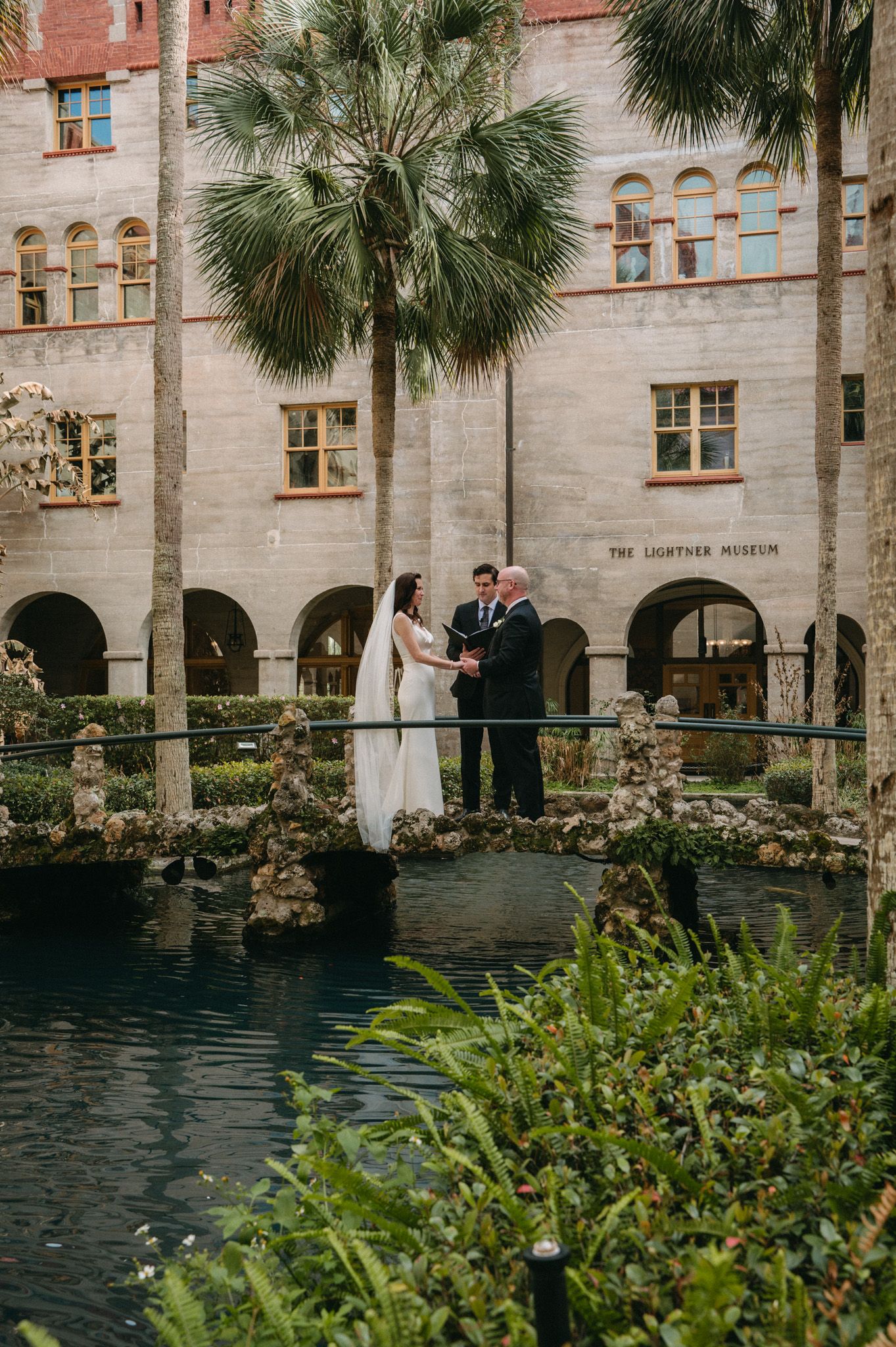 Couple exchanges vows on a bridge over a pond at the Lightner Museum in St. Augustine, FL, surrounded by palm trees.